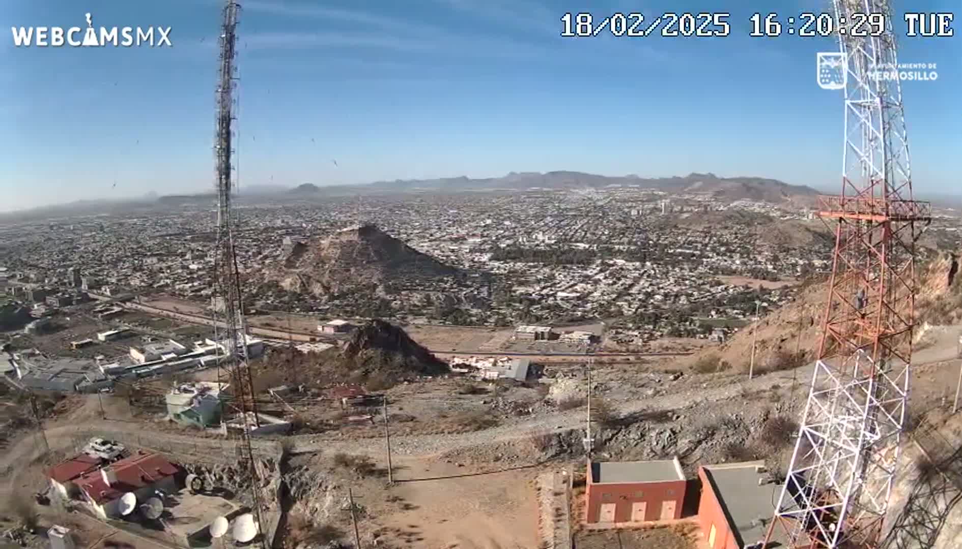 A high-angle, long shot on a clear day shows a sprawling city below a rocky hilltop with communication towers, buildings, and satellite dishes.