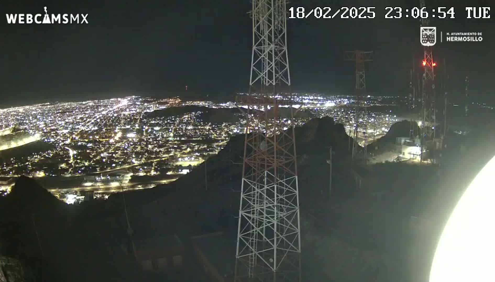 A nighttime view from a hilltop overlooking a brightly lit city, with two tall communication towers in the foreground.