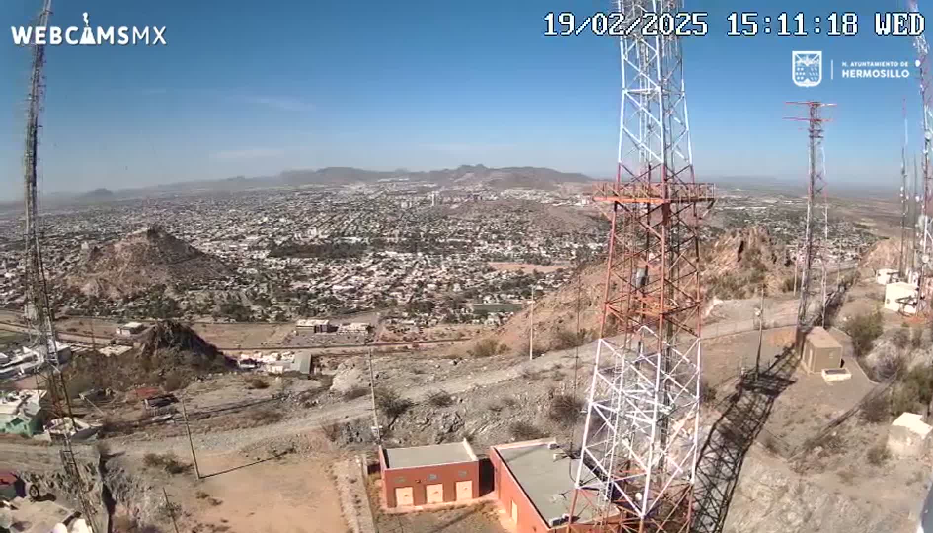 A high-angle, sunny view from a hilltop shows a sprawling city extending to distant mountains, with several tall communication towers in the immediate foreground.
