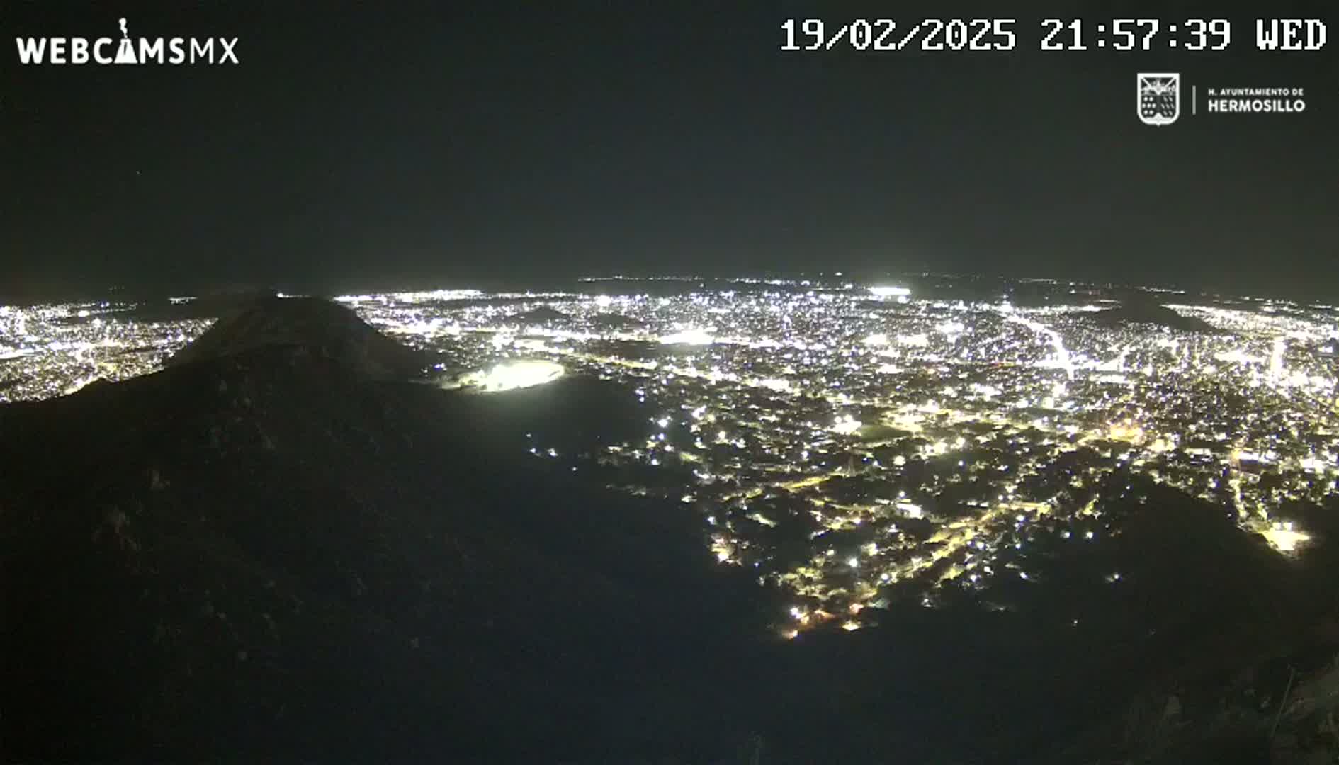 A nighttime, high-angle view shows a sprawling city illuminated below a dark, silhouetted hill.