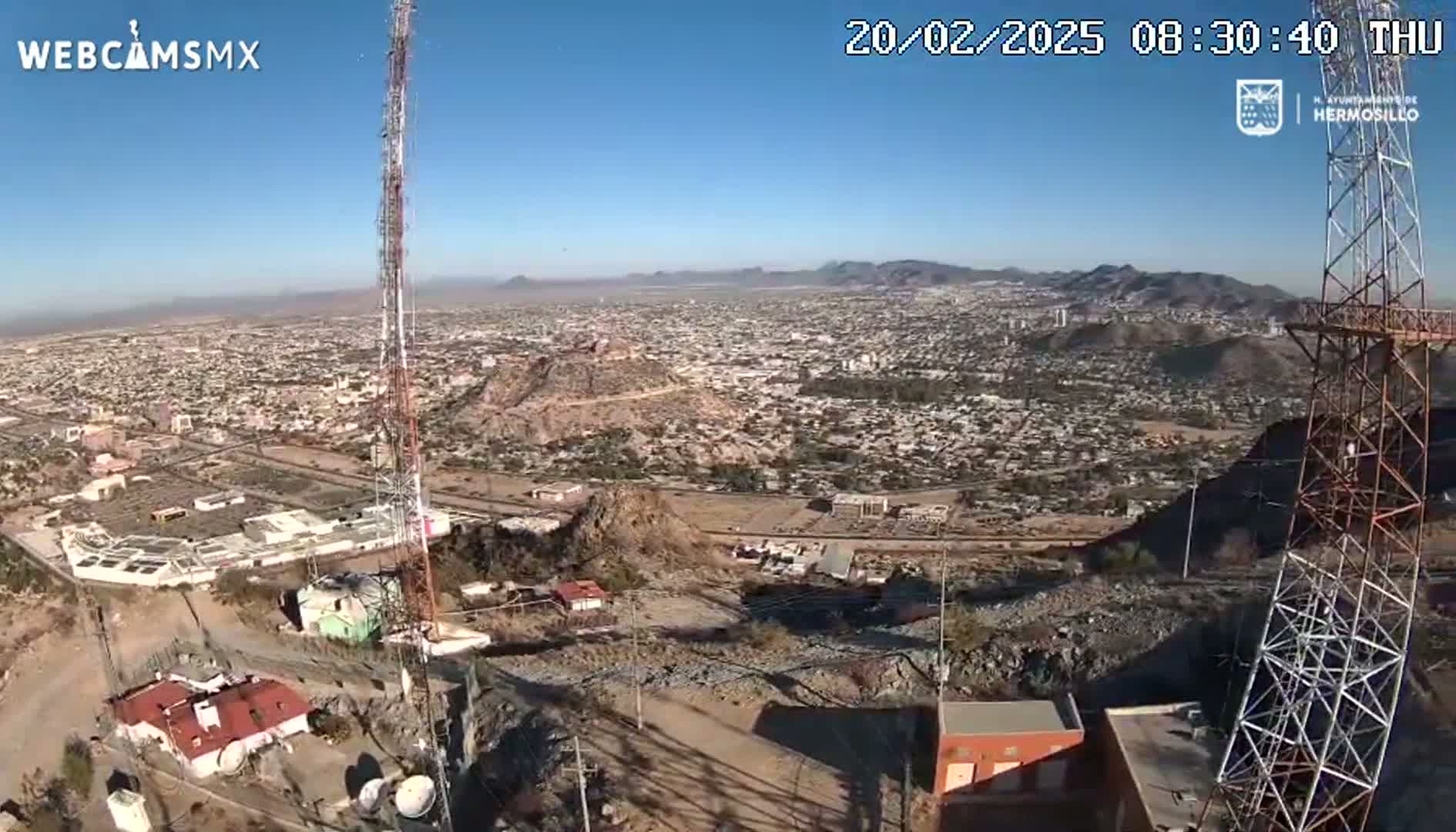 A high-angle, long shot on a clear day shows a sprawling city nestled in a mountainous region, with two tall communication towers prominently positioned in the foreground.