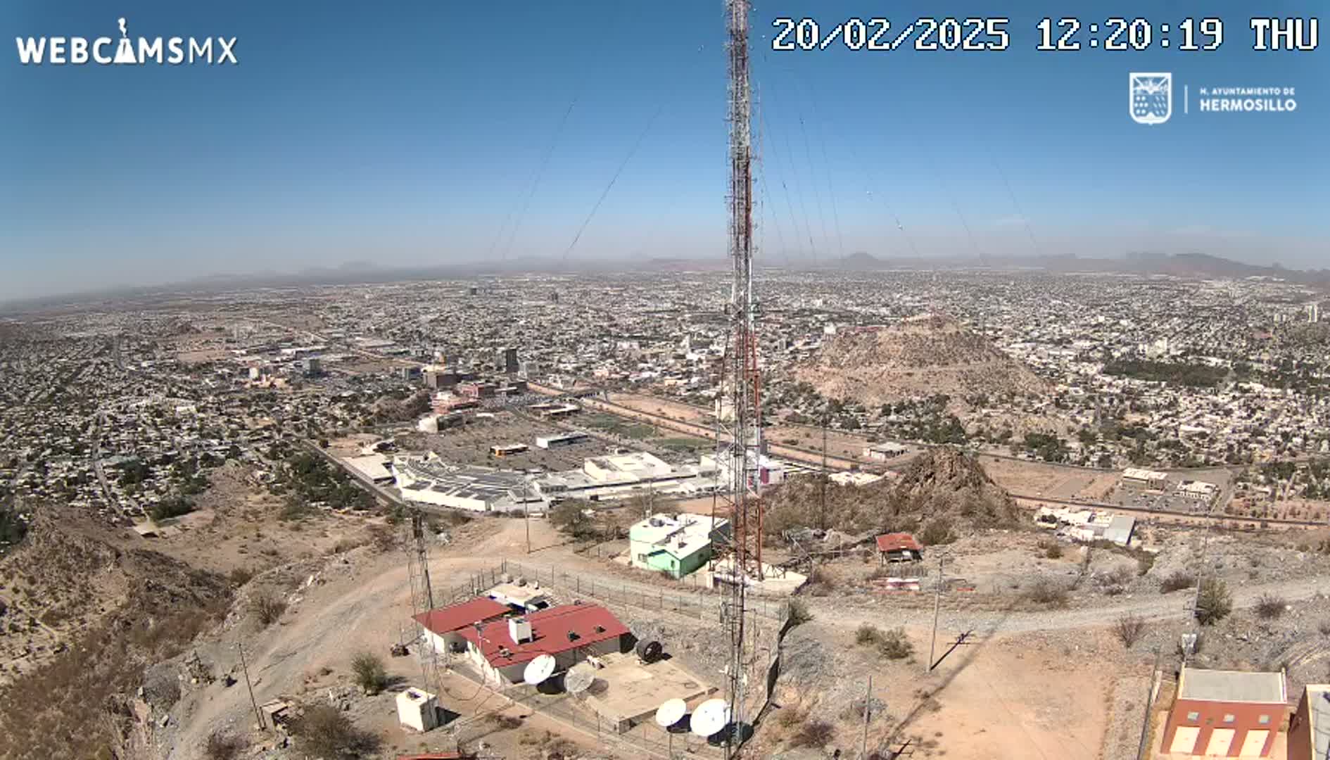 A high-angle, long shot on a clear, sunny day shows a large city sprawling across a desert landscape, with a tall communication tower and several buildings situated on a rocky hillside overlooking it.