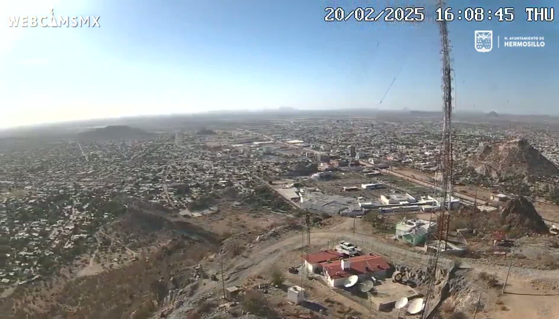 A high-angle, long shot on a clear, sunny day shows a sprawling city extending to the horizon, with rocky hills and a communications tower in the foreground.