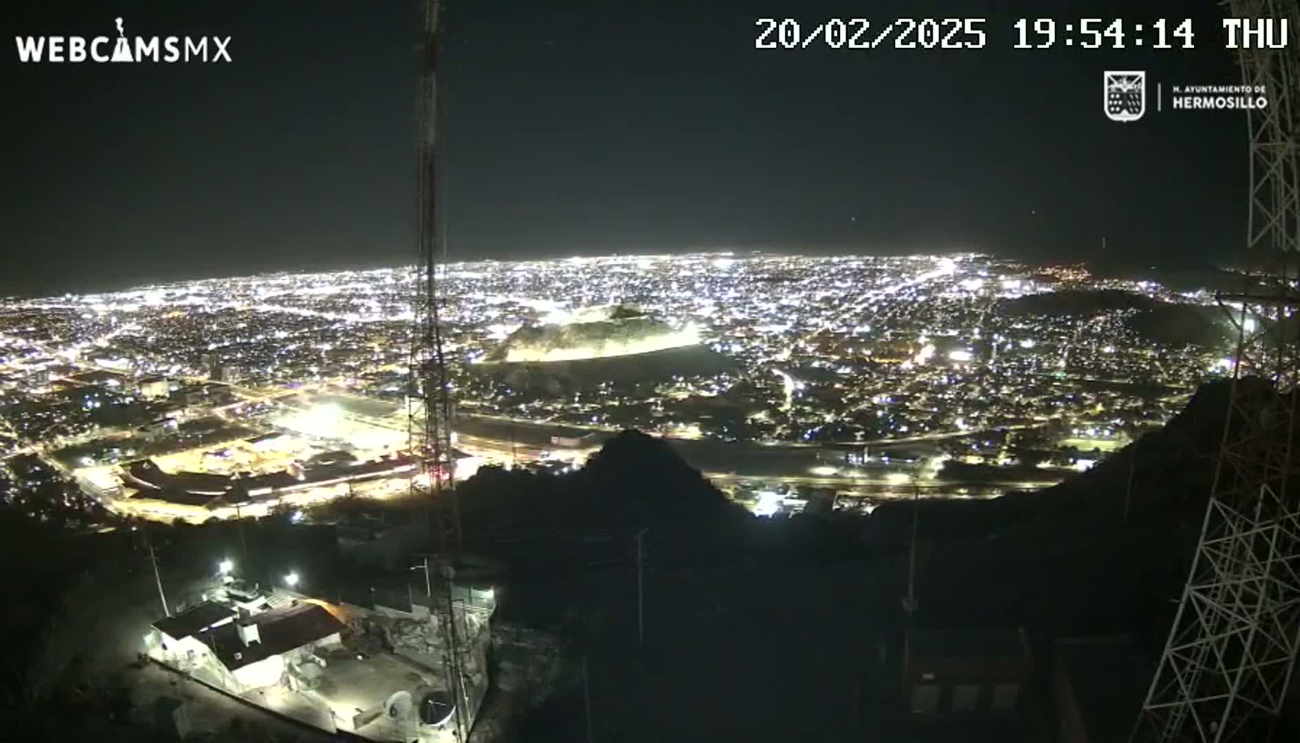 A nighttime, aerial view shows a sprawling city illuminated below, seen from a vantage point on a hill with communication towers.