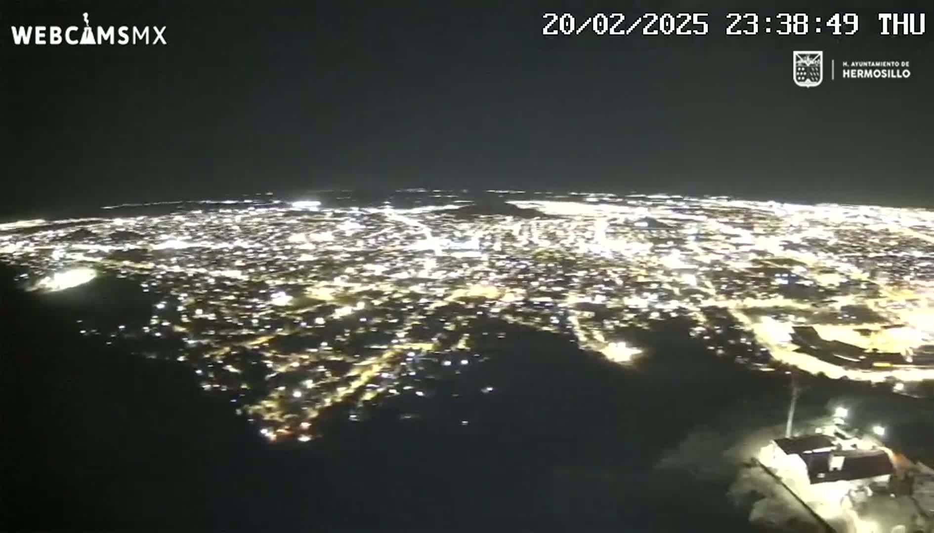 A nighttime aerial view shows a sprawling city illuminated by countless lights, with a small building visible in the bottom right corner under a clear, dark sky.