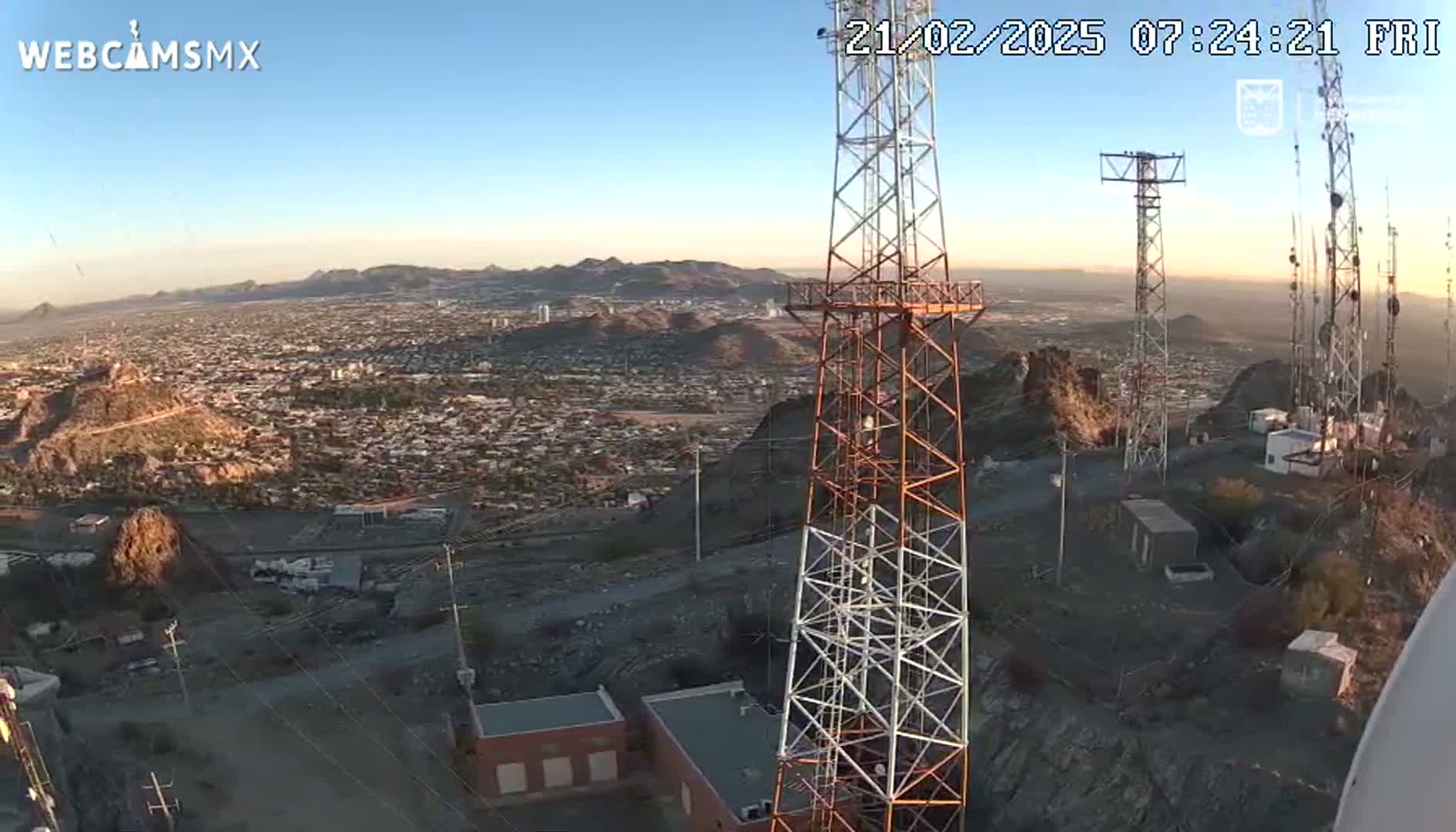 A high-angle, long shot of a city sprawling below a hilltop with several communication towers on a clear, sunny day.