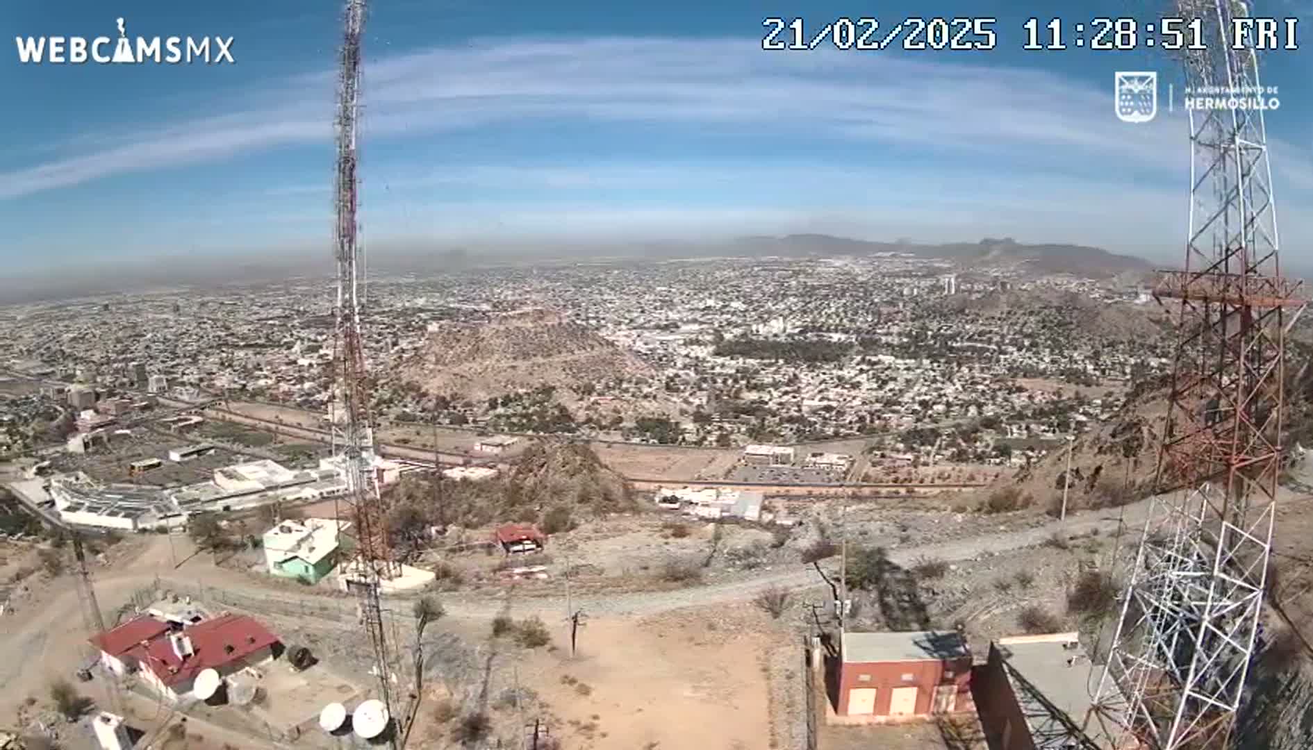 A sprawling city is visible under a mostly clear, sunny sky, seen from a high vantage point near tall communication towers and buildings.
