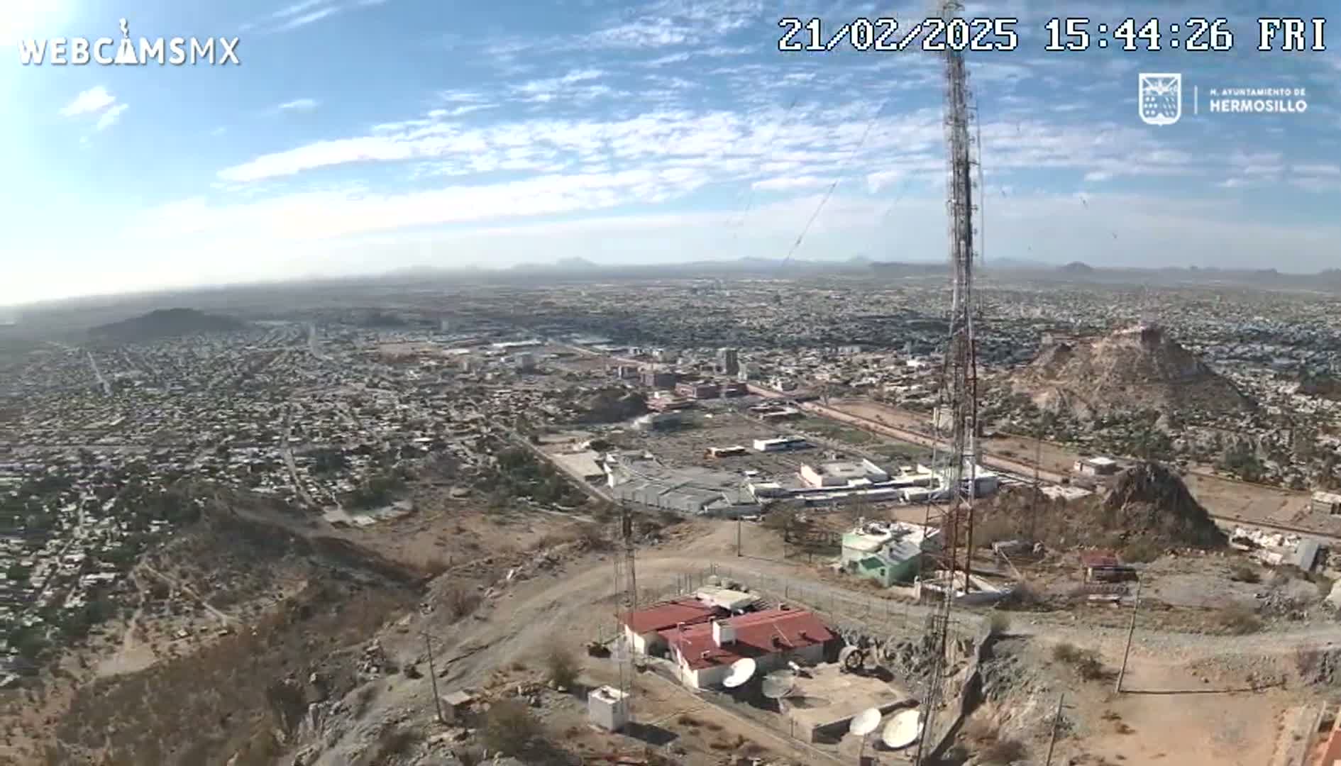 A high-angle, long shot on a clear, sunny day shows a sprawling city nestled below a hilltop, where a small building and tall communication tower are situated.