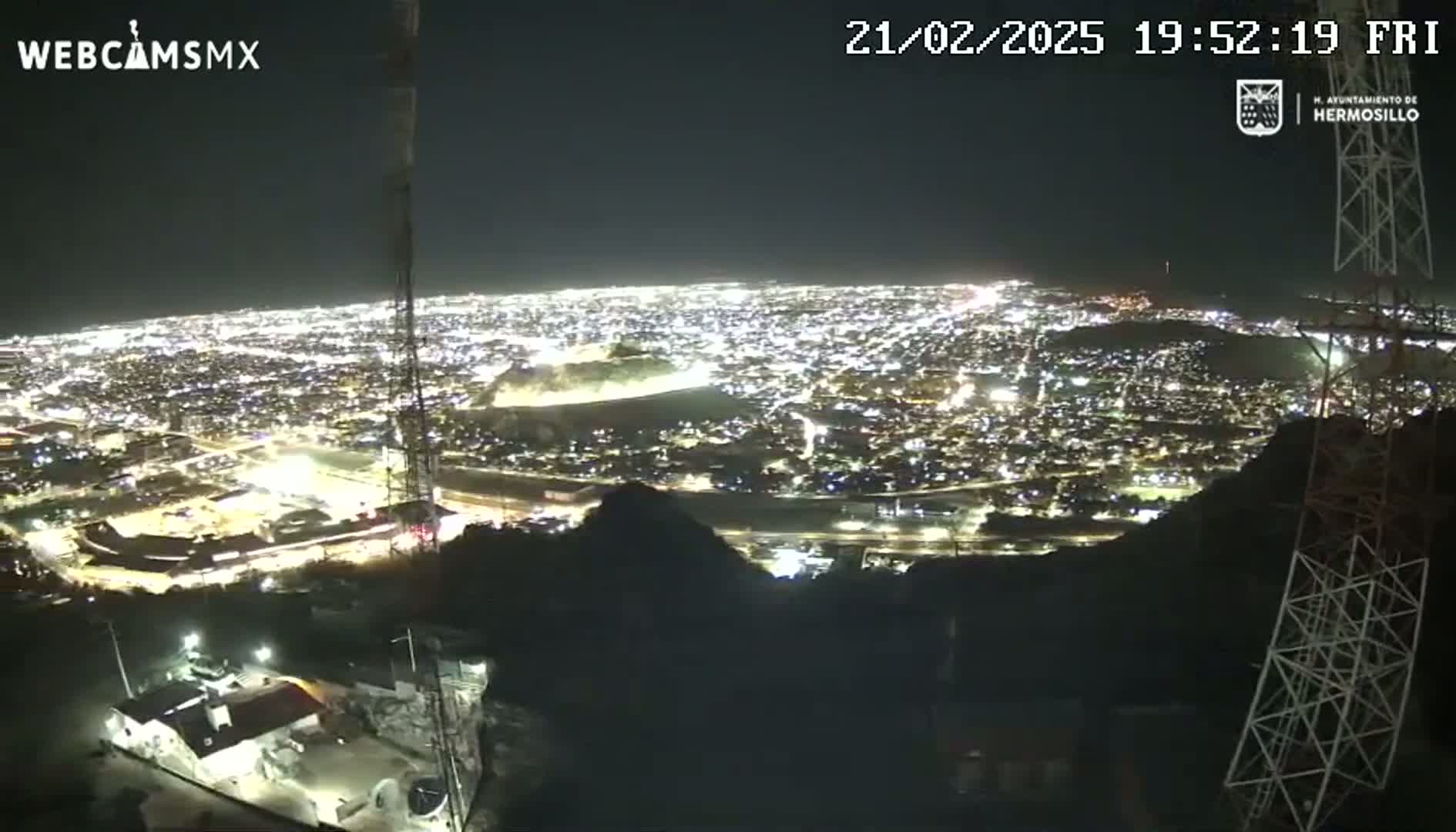 A nighttime, high-angle view shows a sprawling city illuminated below, seen from a vantage point on a hilltop with communication towers present.