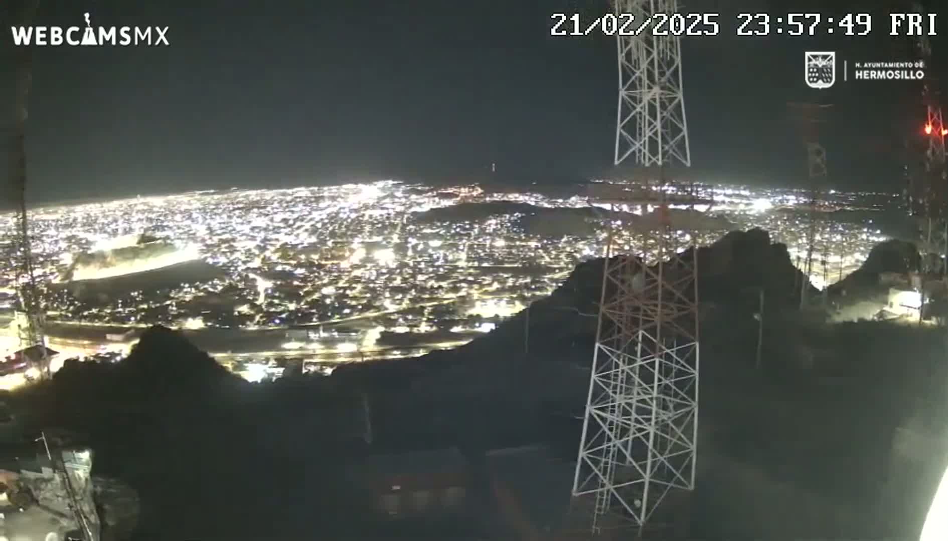 A nighttime view from a hilltop shows a sprawling city illuminated below, with two tall communication towers in the foreground under a clear sky.