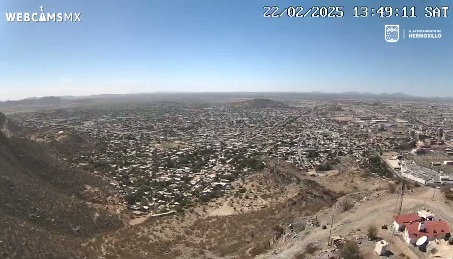 A sprawling city is seen from a high vantage point on a clear, sunny day, with arid, brown hills in the foreground.