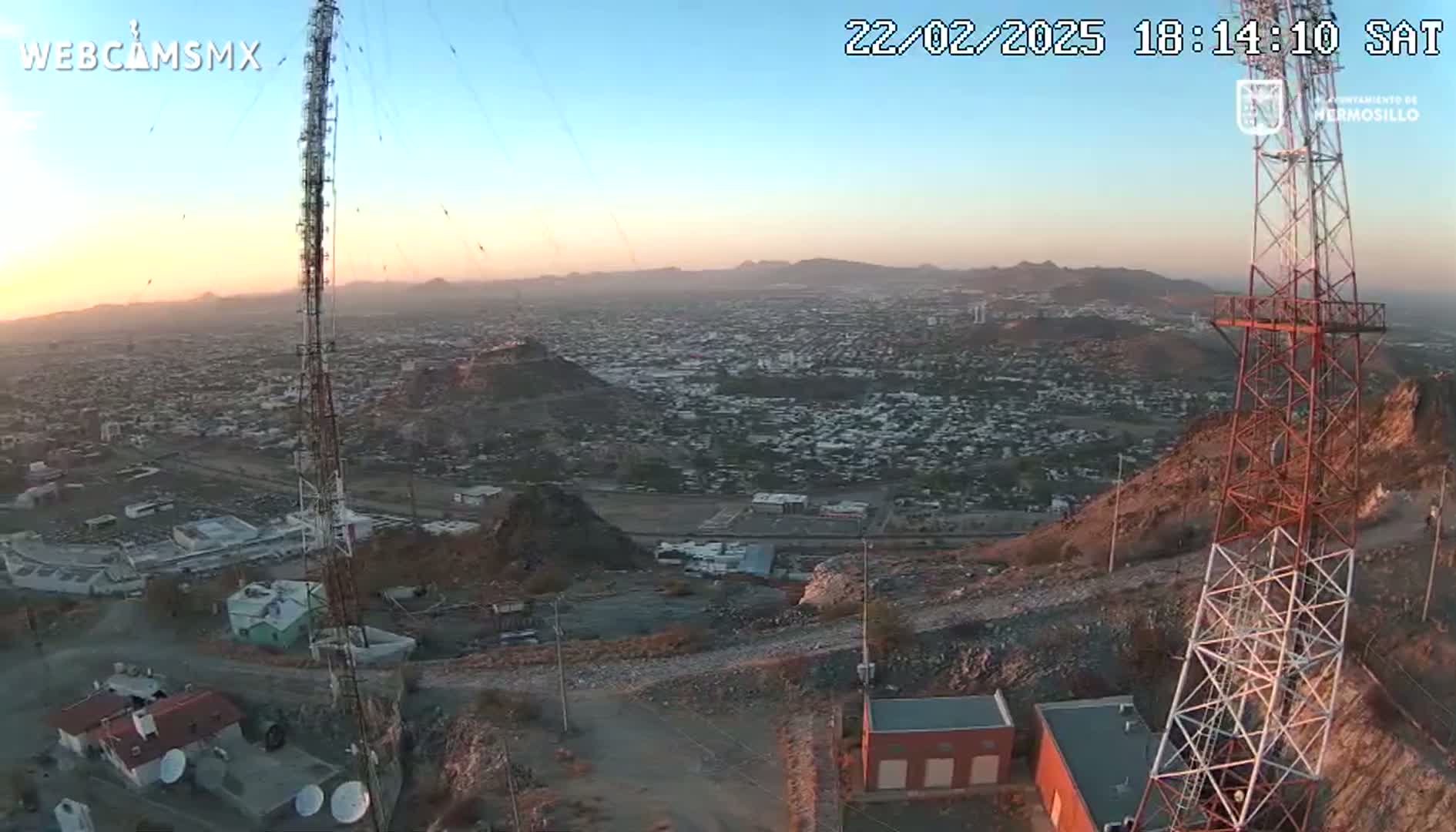 A sprawling city is visible at sunset from a hilltop, where two communication towers stand near some small buildings.