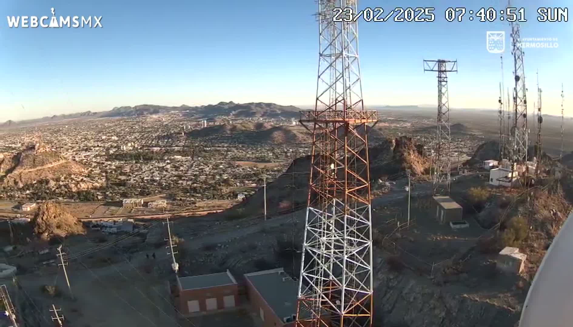 A sprawling city is visible beneath a clear sky from a high vantage point on a rocky hilltop, near several tall communication towers.
