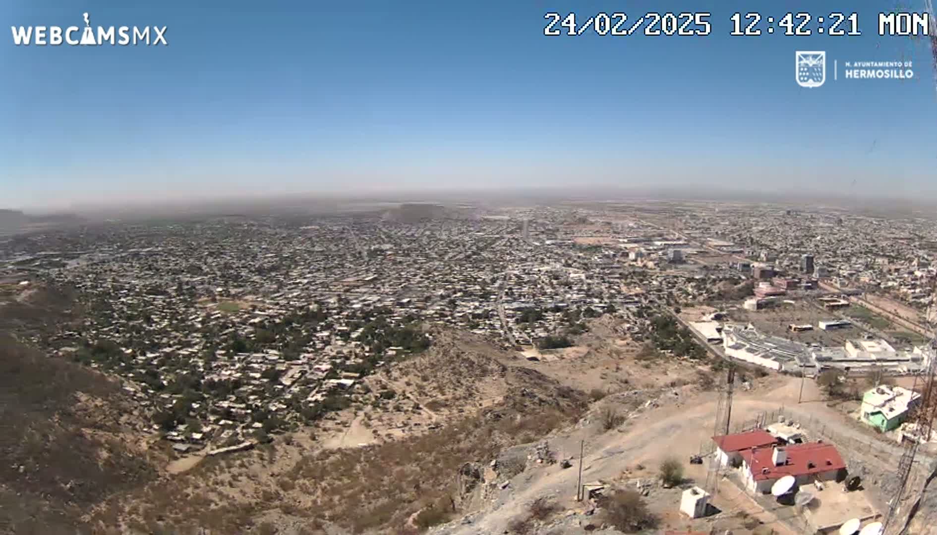 A sprawling city is viewed from a high vantage point on a hazy, sunny day, with a rocky, sparsely vegetated hillside in the foreground.