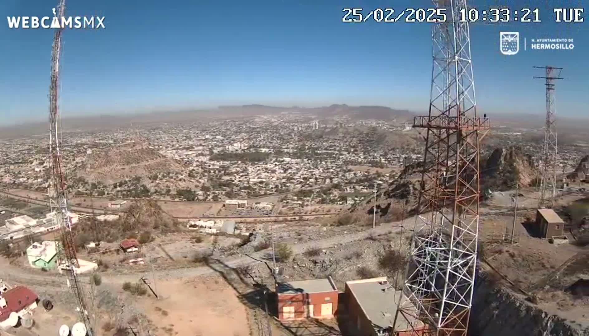 A high-angle, long shot on a clear, sunny day shows a sprawling city below two tall communication towers perched atop a rocky hill.