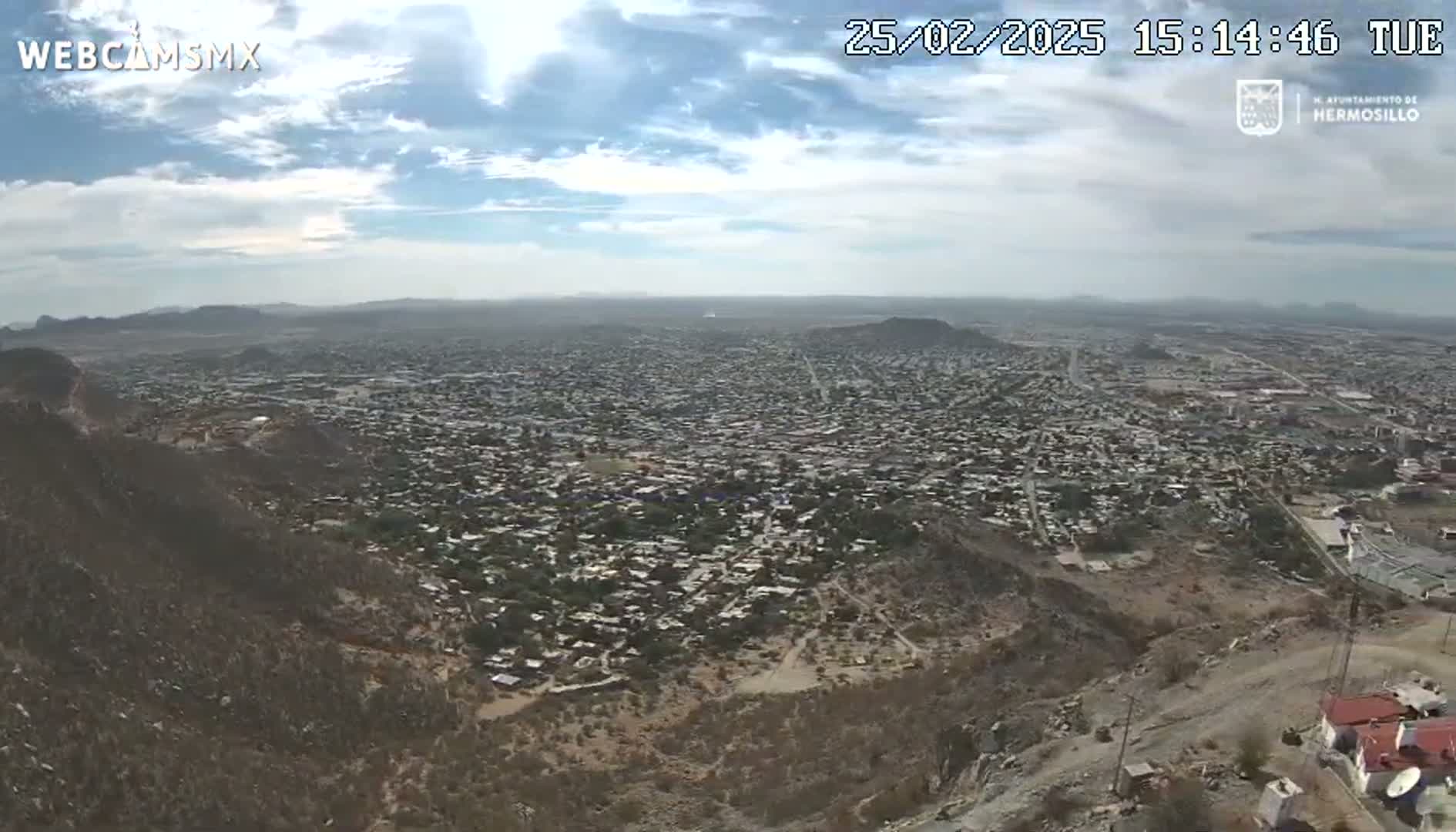 A sprawling city is viewed from a high vantage point on a partly cloudy day, with arid hillsides in the foreground.