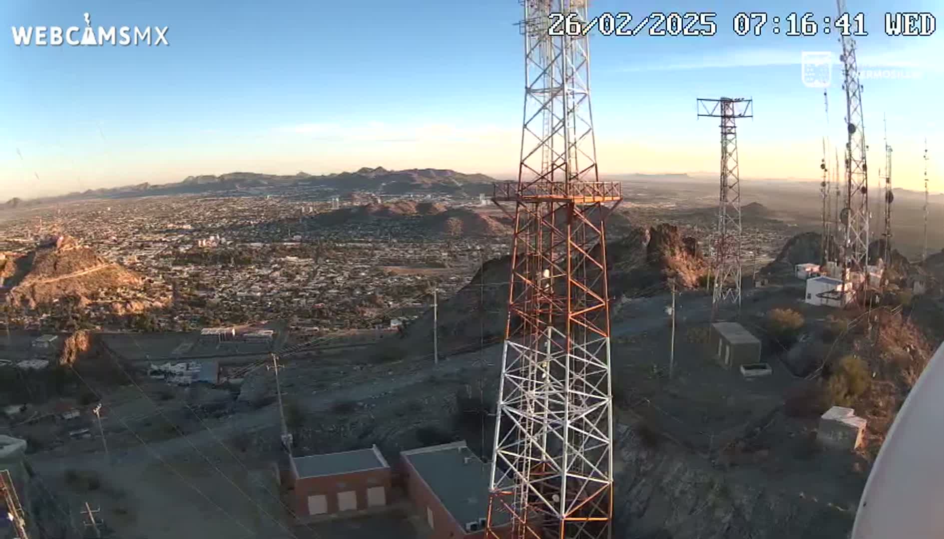 A sprawling city is visible below several communication towers on a hilltop at sunrise under a clear sky.