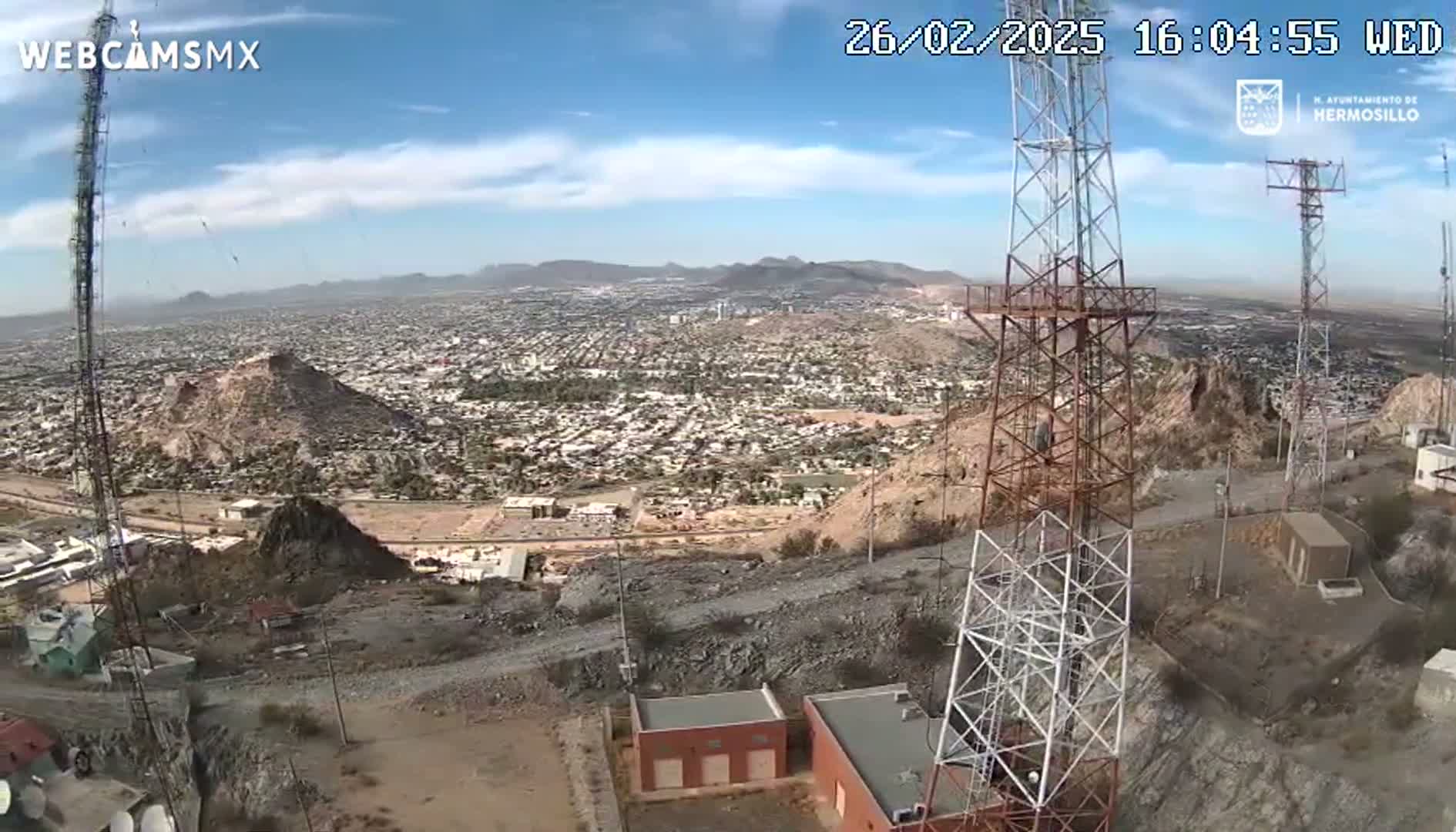 A high-angle, daytime view shows a sprawling city nestled among hills and mountains under a mostly clear sky, with two tall communication towers prominent in the foreground.