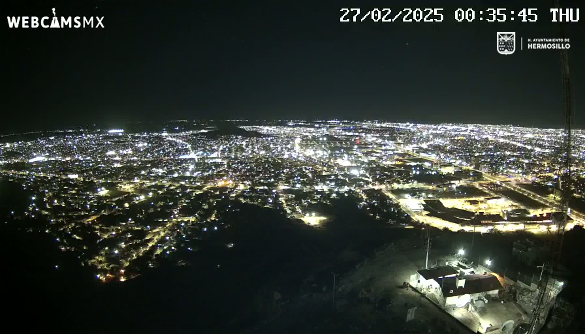 A nighttime, high-angle view shows a sprawling city illuminated by countless lights, with clear skies.