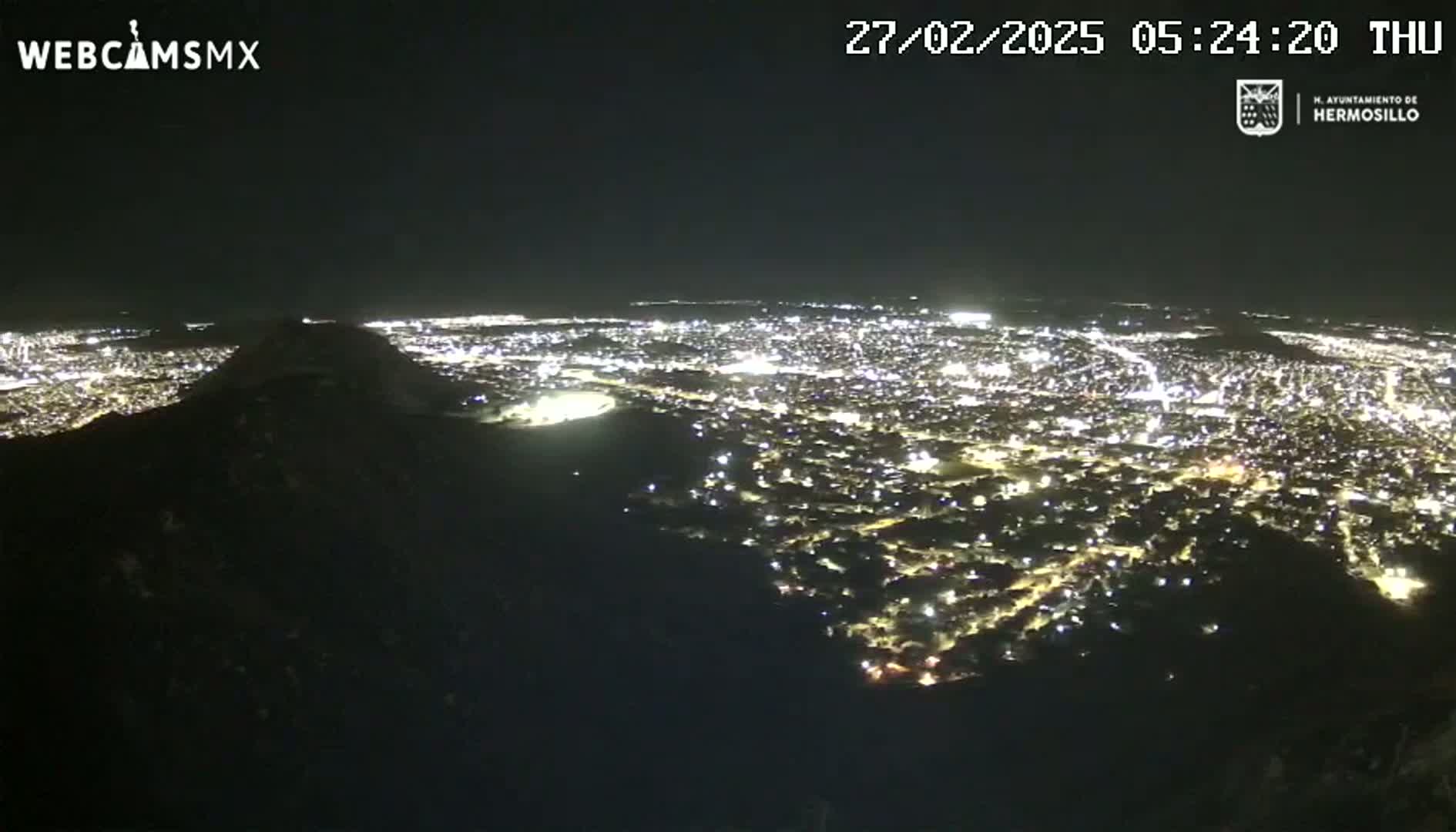 A nighttime, high-angle view shows a sprawling city illuminated by numerous lights, partially obscured by a dark hill in the foreground.