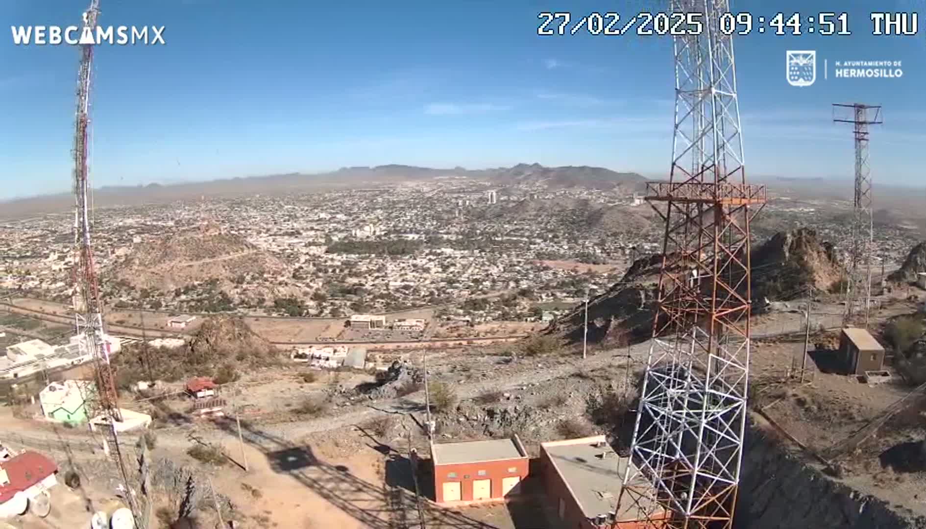 A high-angle, long shot on a clear, sunny day shows a sprawling city nestled in a mountainous region, with two tall communication towers prominently featured in the foreground.