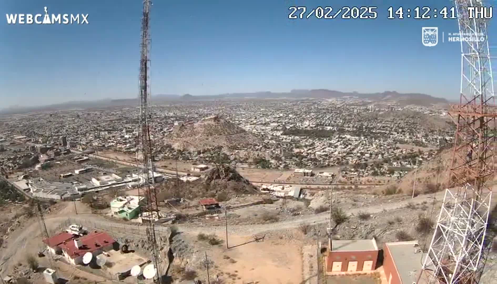 A high-angle, long shot on a clear, sunny day shows a sprawling city below a hilltop, with communication towers and buildings on the hill.