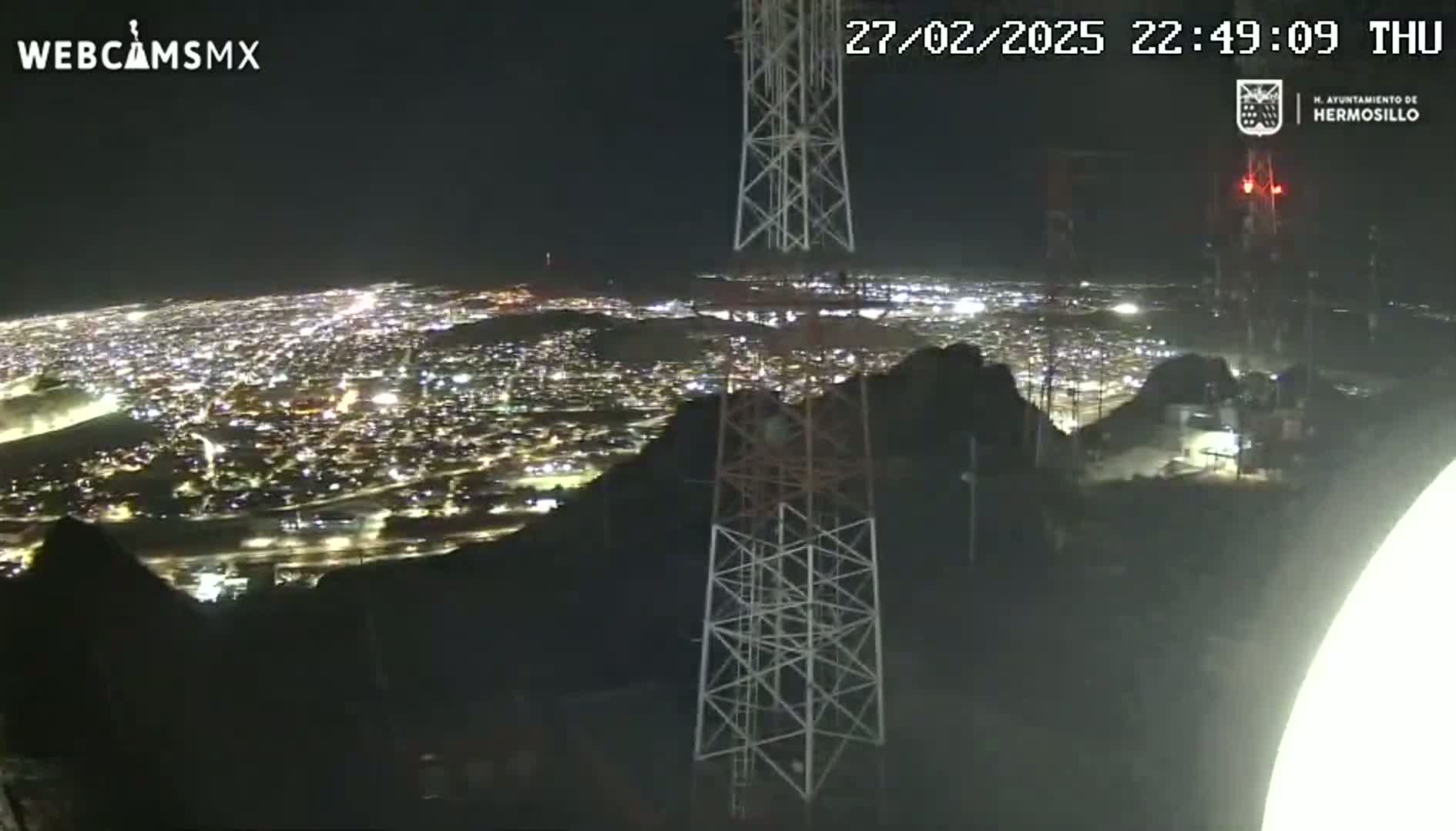 A nighttime, high-angle view shows a sprawling city illuminated below, nestled between dark hills and overlooked by several tall communication towers.