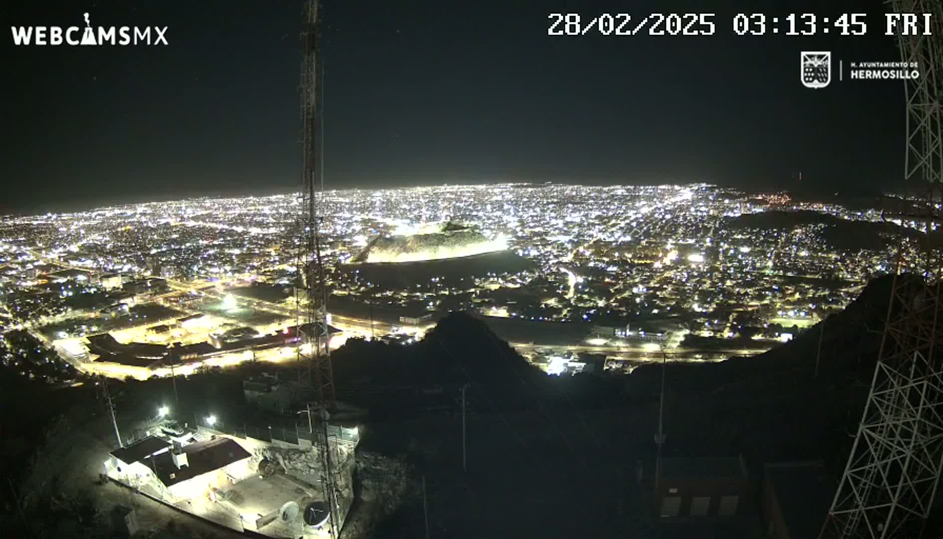 A nighttime, high-angle view from a hilltop shows a sprawling city illuminated by many lights.