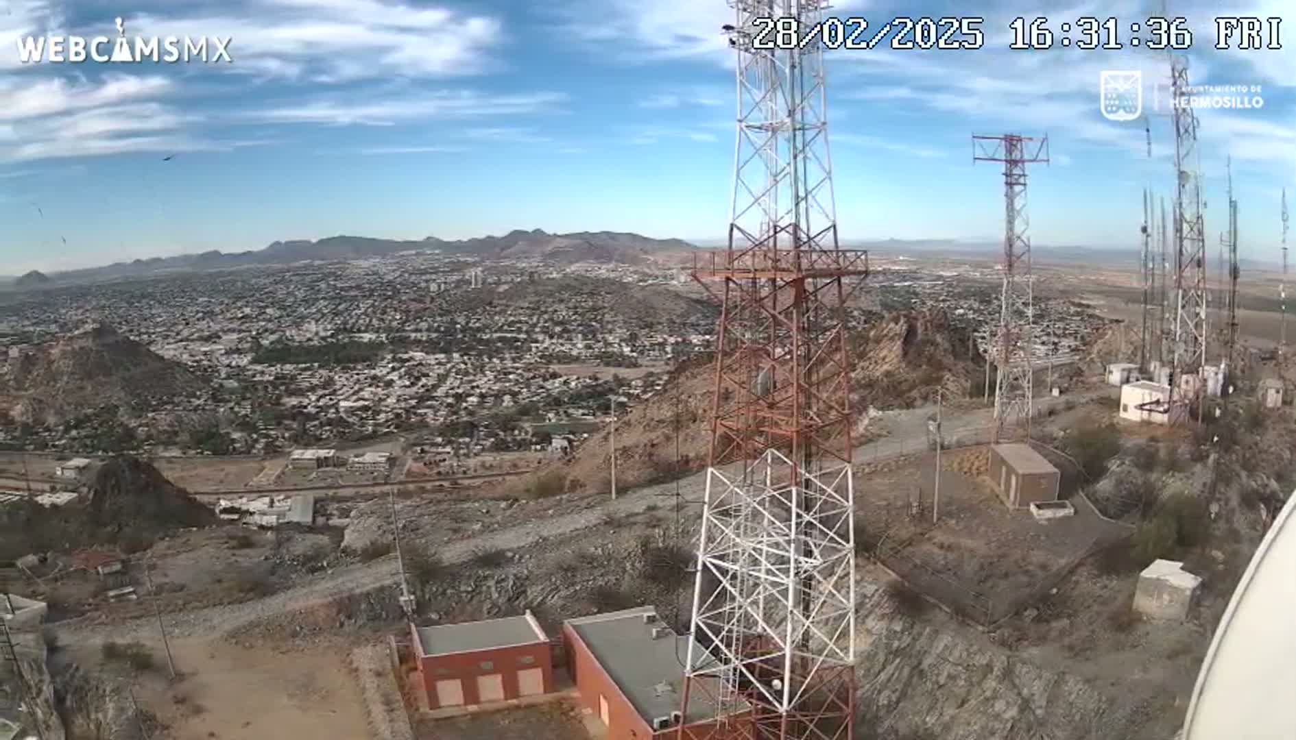 A high-angle view on a partly sunny day shows several communication towers atop a rocky hill overlooking a sprawling city in a desert or arid environment.