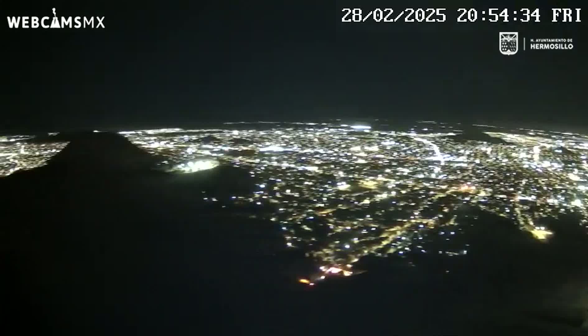 A nighttime, high-angle view shows a sprawling city illuminated by numerous lights, partially obscured by a dark mountain or hill in the foreground.