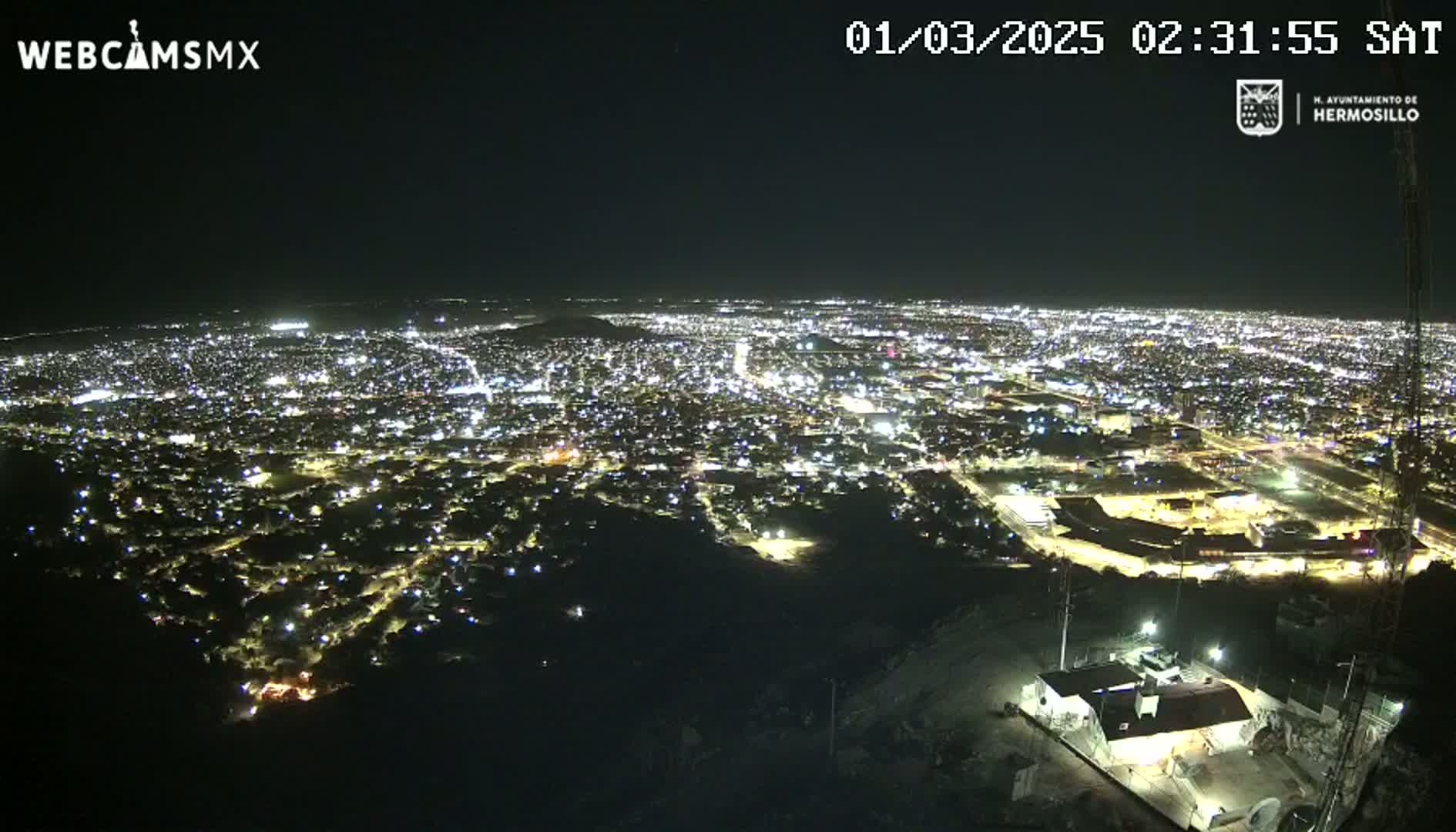 A nighttime, high-angle view shows a sprawling city illuminated by countless lights, extending to the horizon under a clear, dark sky.