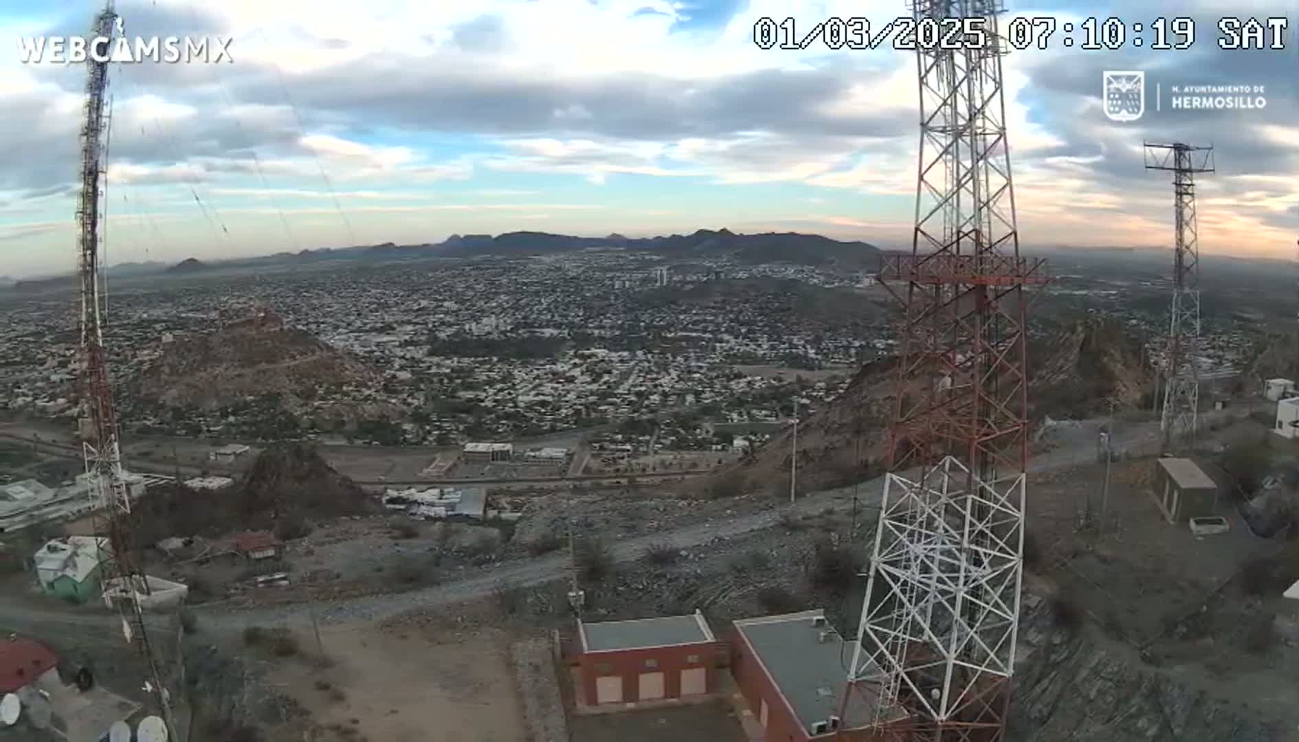 A high-angle, long shot shows two tall communication towers on a hill overlooking a sprawling city under a partly cloudy sky.