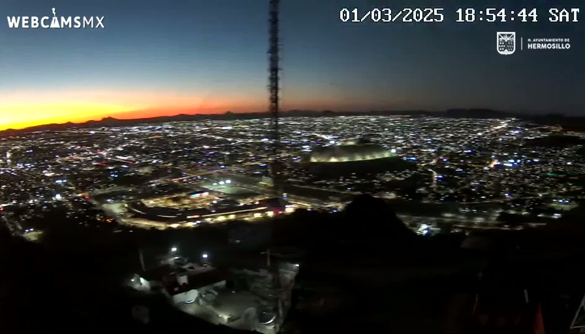 A nighttime, high-angle view of a city illuminated by lights, with a sunset visible in the distance.