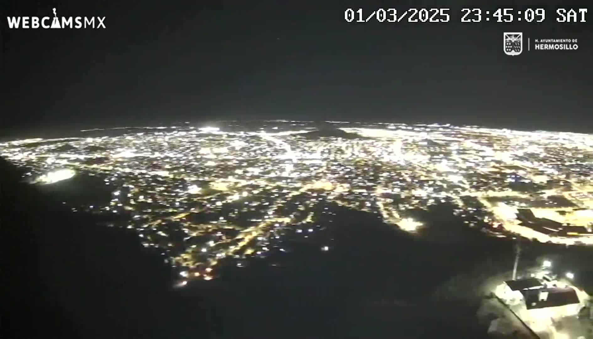 A nighttime, high-angle view shows a sprawling city illuminated by countless lights, stretching across a dark landscape.