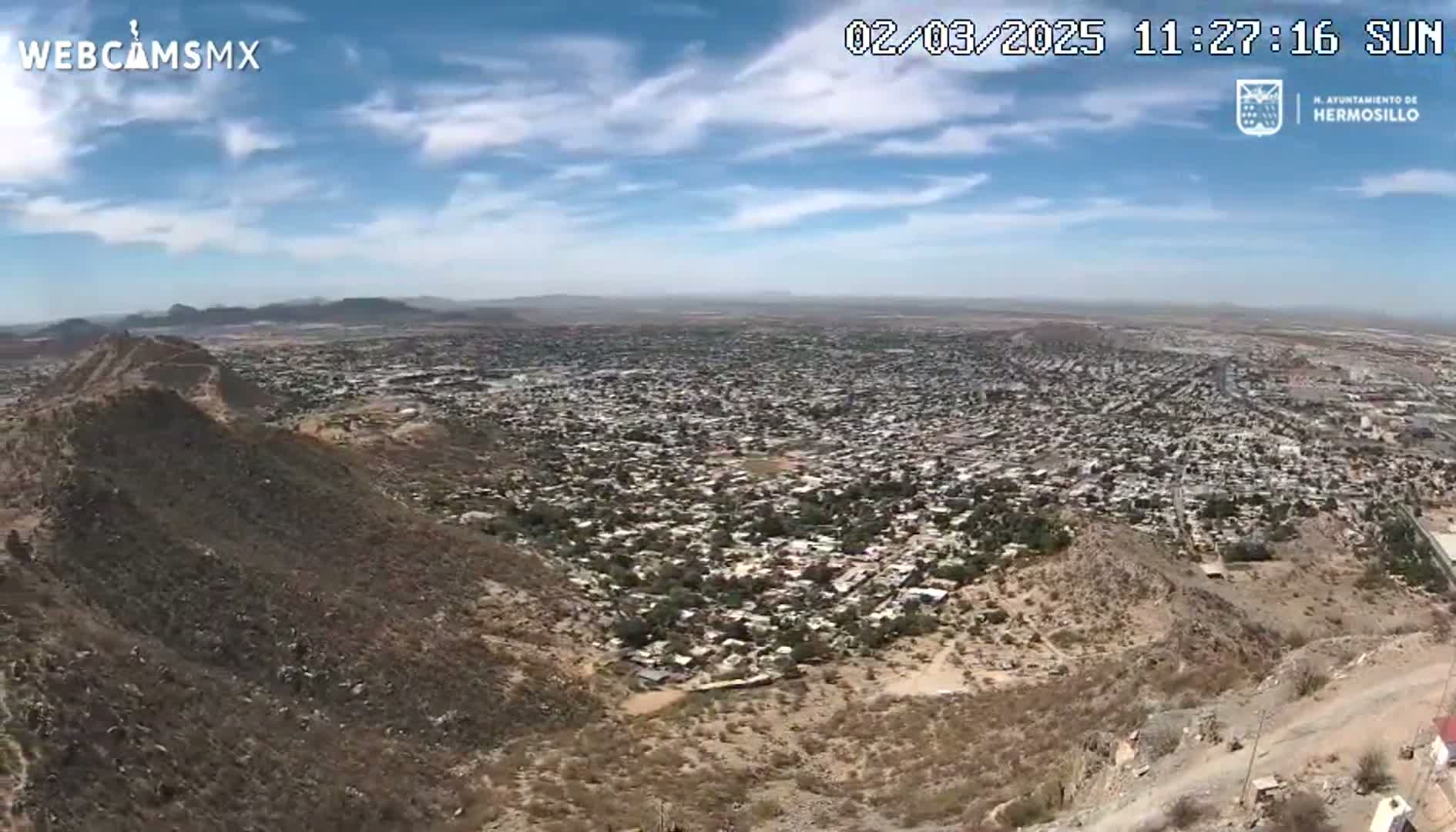 A sprawling city is visible from a high vantage point on a sunny day with few clouds, nestled among arid, brown hills.
