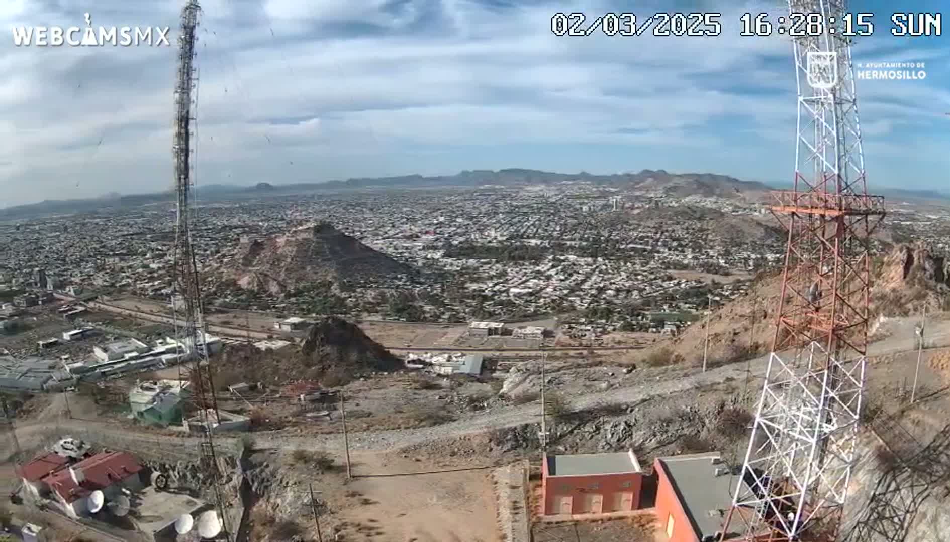 A high-angle, sunny view shows a large city sprawling across a valley, with a rocky, arid hillside in the foreground containing communication towers and small buildings.