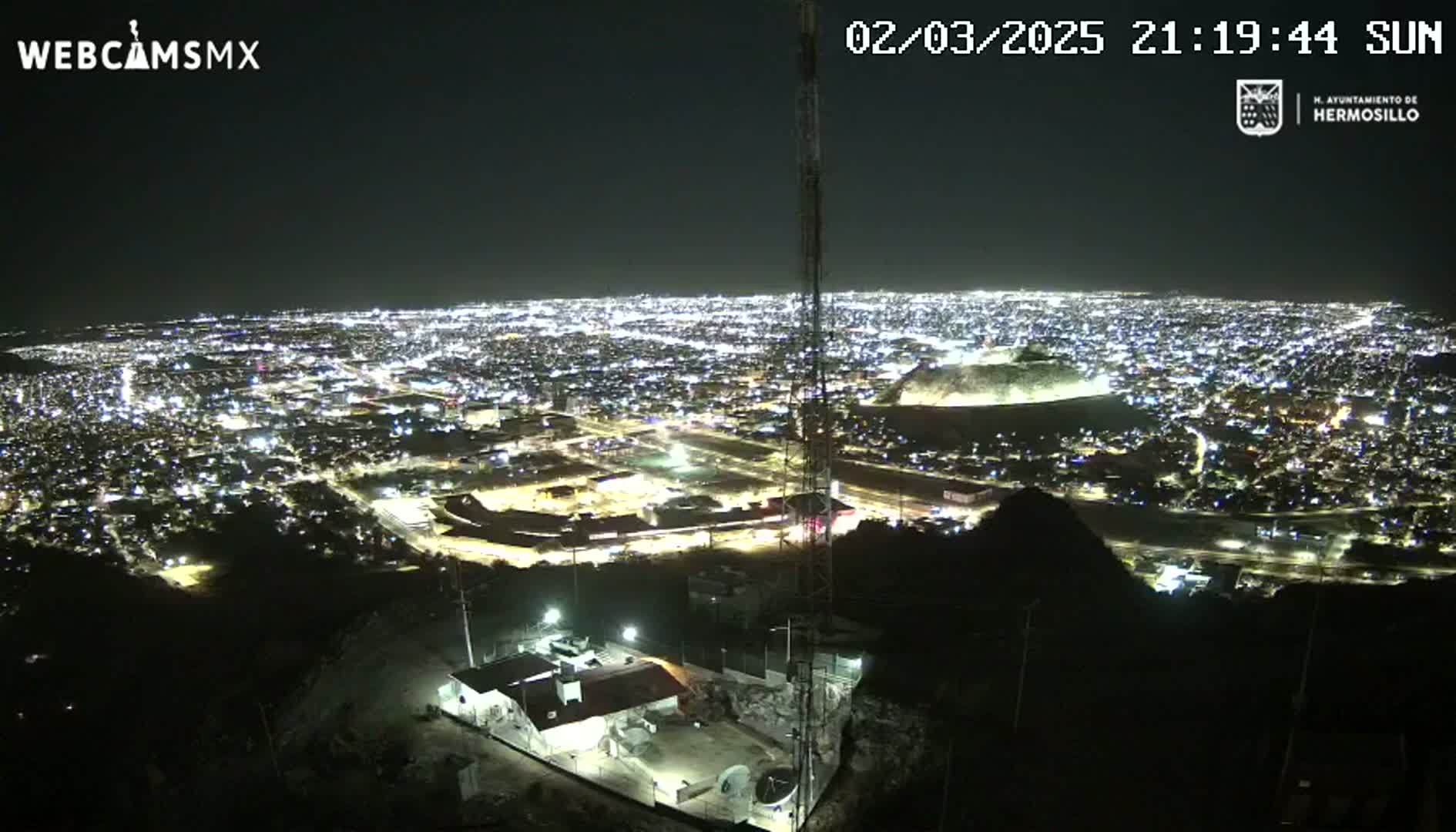A nighttime aerial view of a brightly lit city sprawling across a dark landscape, seen from a hilltop vantage point.