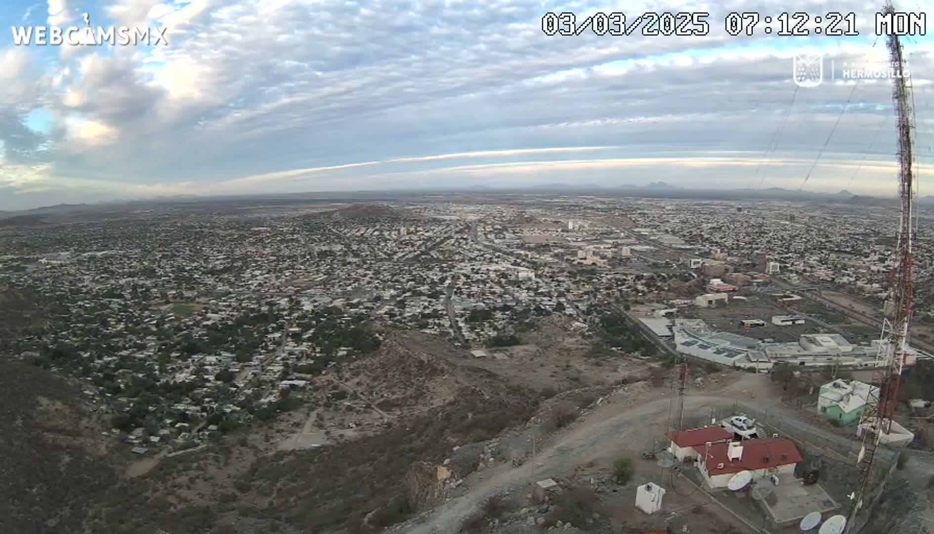 A high-angle view of a sprawling city under a partly cloudy sky, seen from a hilltop with communication towers and buildings in the foreground.