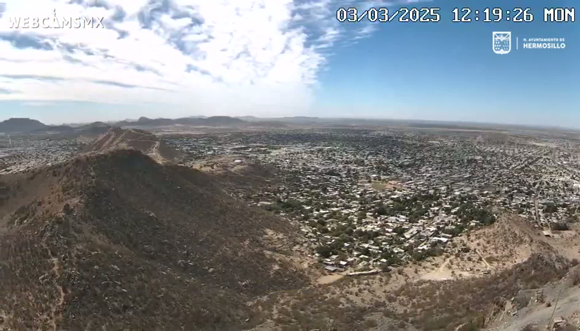 A sprawling city is visible below from a high vantage point on a partly cloudy day, with hills and mountains in the surrounding area.