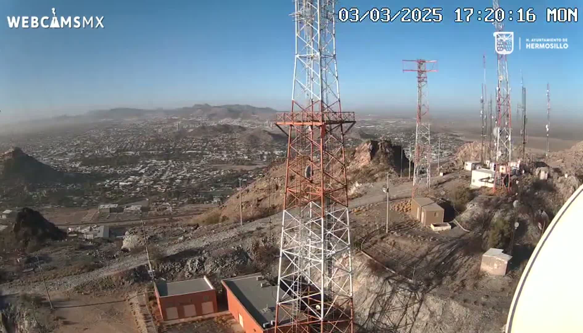 A high-angle view on a clear day shows several communication towers atop a rocky hill overlooking a sprawling city.