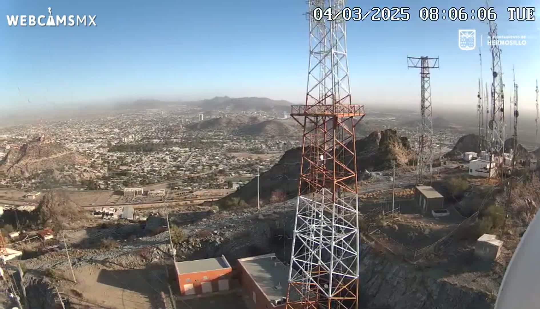 A hazy view of a city sprawls beneath several communication towers on a rocky hilltop.