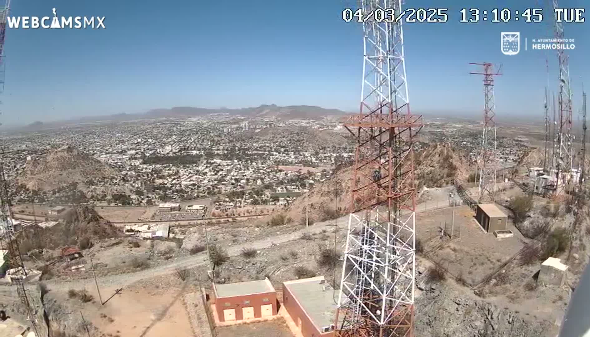 A high-angle, long shot on a clear, sunny day shows a sprawling city viewed from a hilltop, where several communication towers and small buildings are clustered.