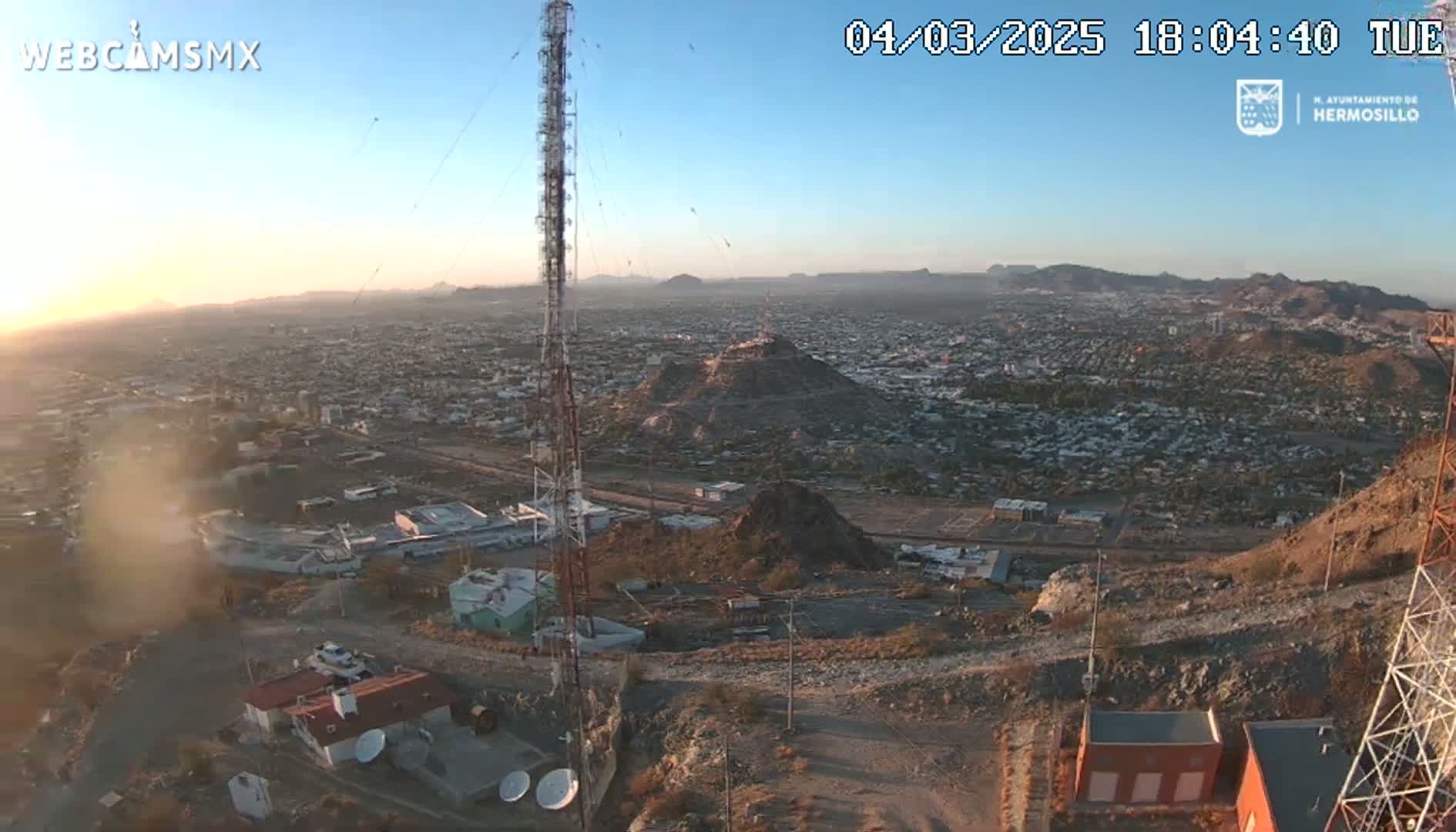 An aerial view at sunset shows a sprawling city nestled in a mountainous region, with communication towers prominent in the foreground.