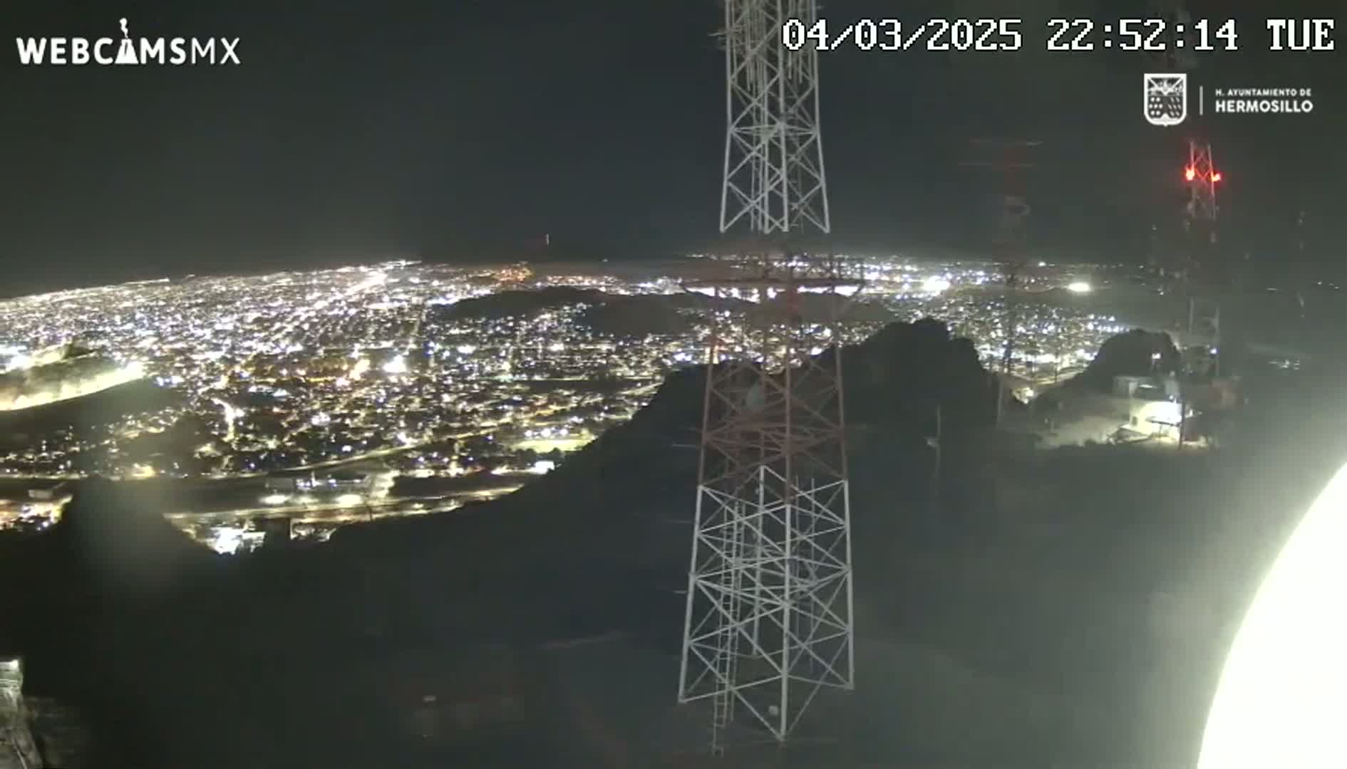 A nighttime view from a hilltop shows a sprawling city illuminated below, with two tall communication towers nearby under a clear sky.