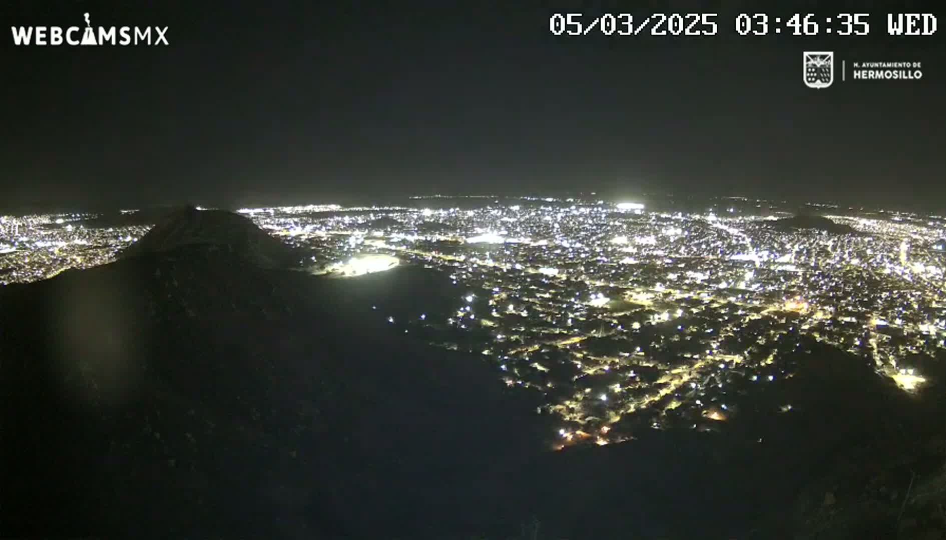 A nighttime, high-angle view shows a sprawling city illuminated by lights, with a dark hill or mountain in the foreground.