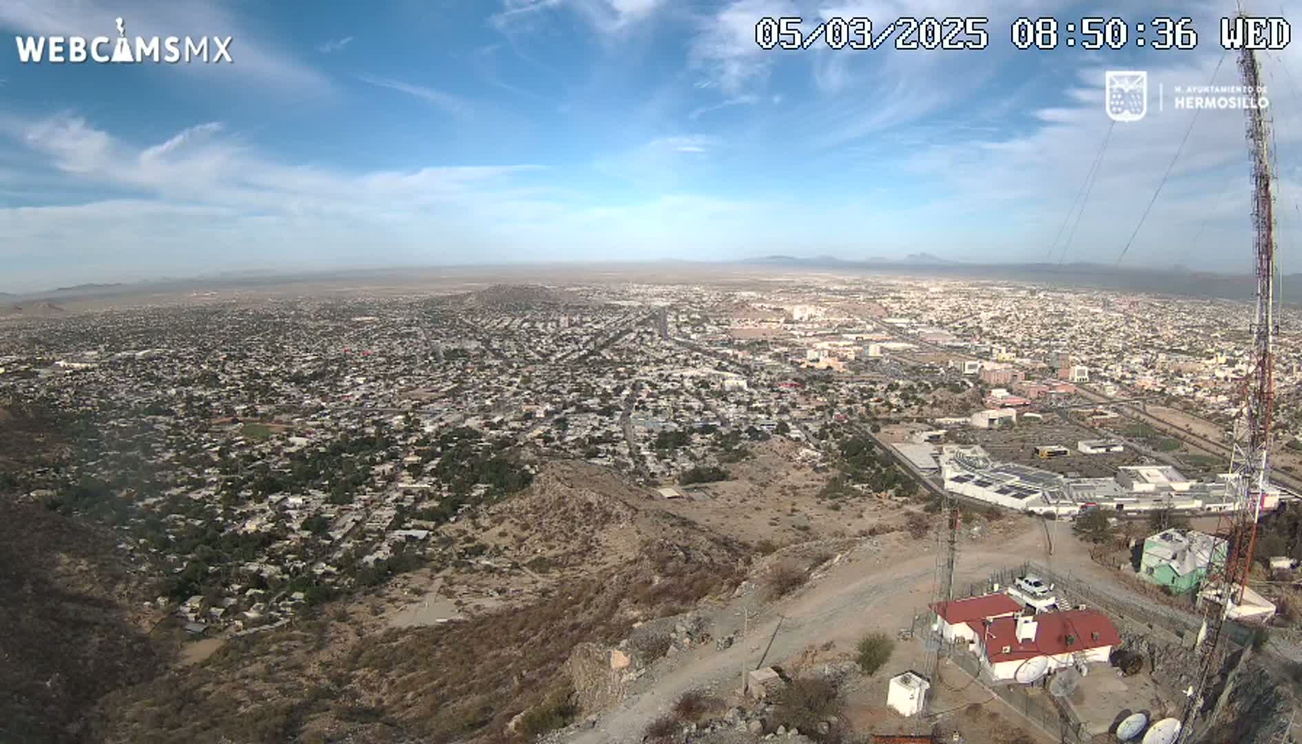 A high-angle, long shot on a clear, sunny day shows a sprawling city nestled between arid hills, with a communications tower and small building visible in the foreground.