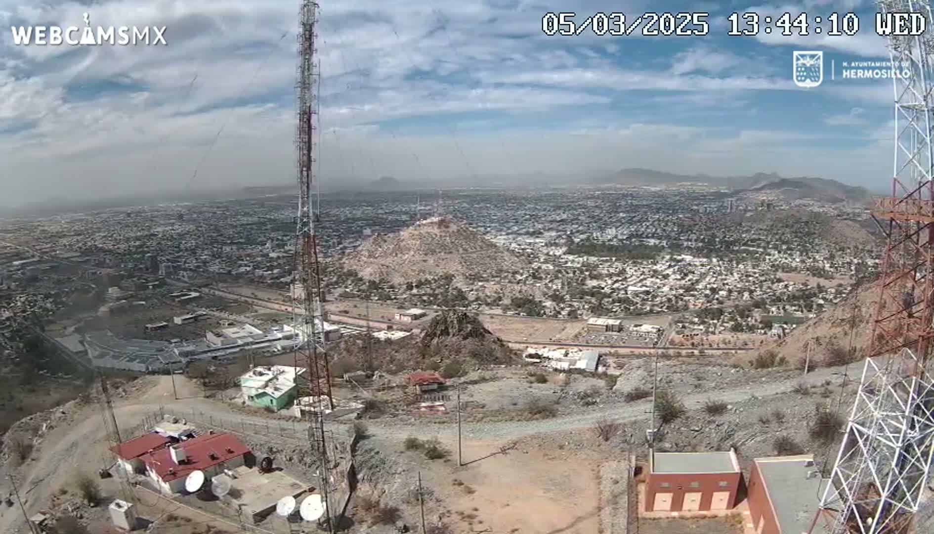 A high-angle view on a partly cloudy day shows a city sprawling below a hilltop with communication towers, buildings, and satellite dishes.