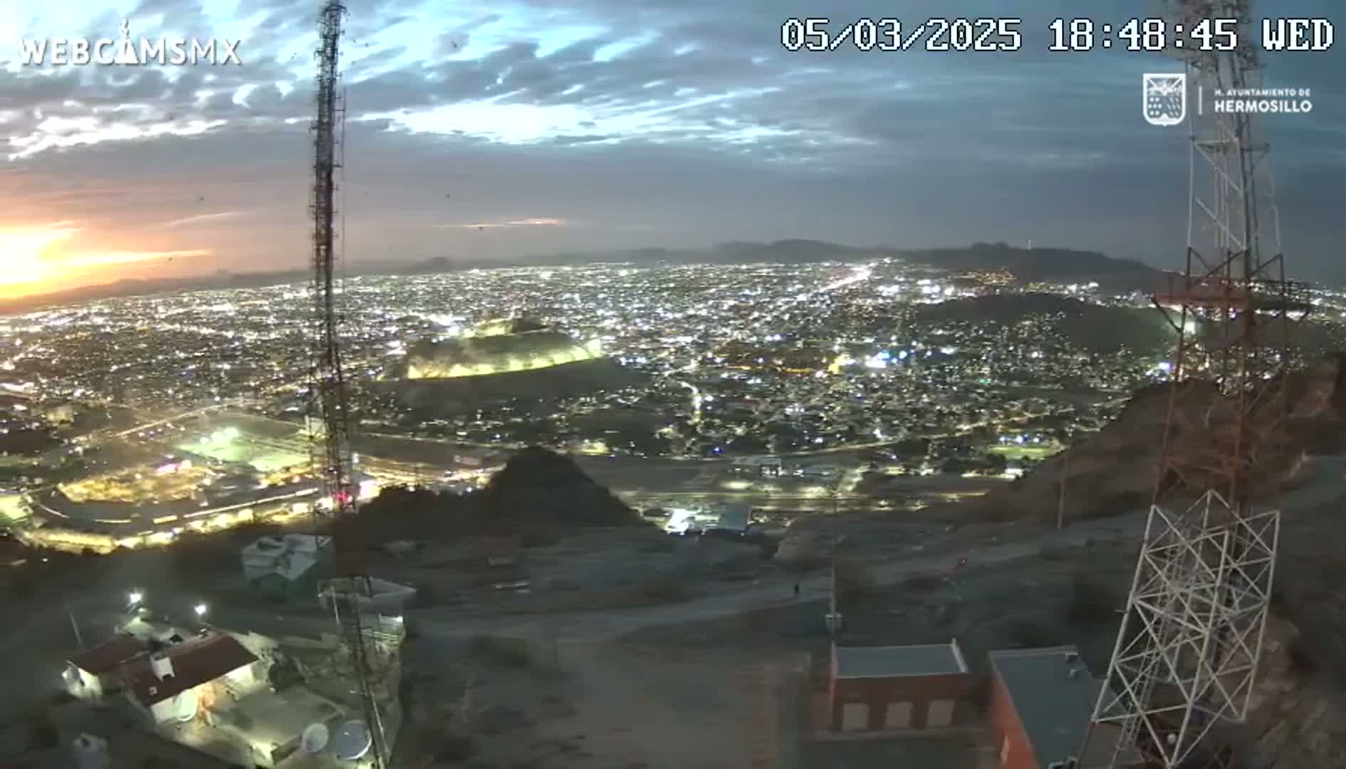 A nighttime view from a hilltop overlooking a brightly lit city with a partly cloudy sky showing the last vestiges of sunset.
