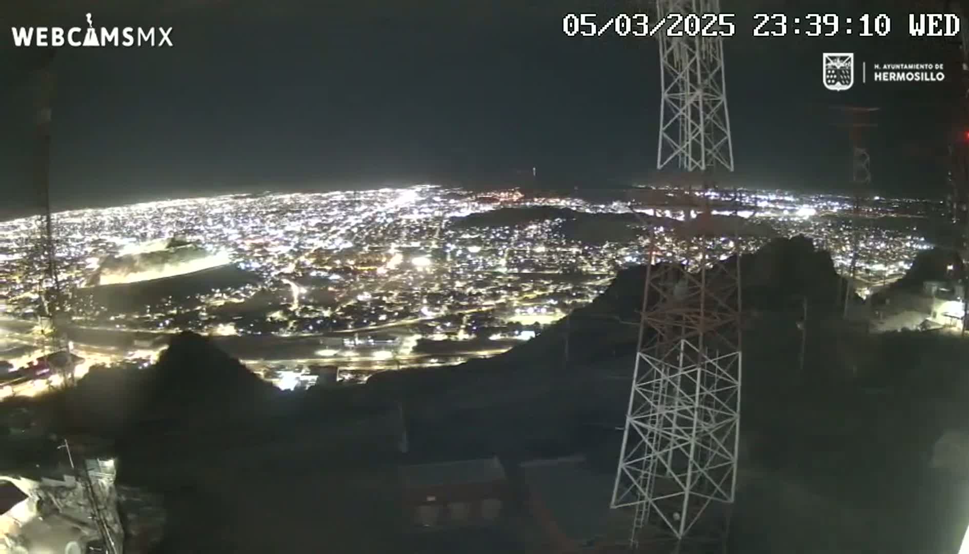 A nighttime, high-angle view shows a sprawling city illuminated by countless lights, seen from a hilltop vantage point near a tall communication tower.