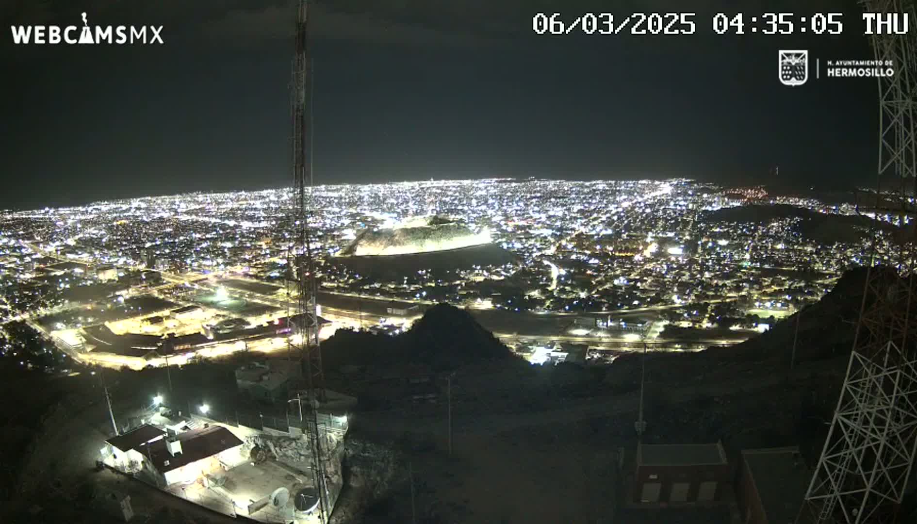 A nighttime, high-angle view shows a sprawling city illuminated by lights, observed from a hilltop vantage point.